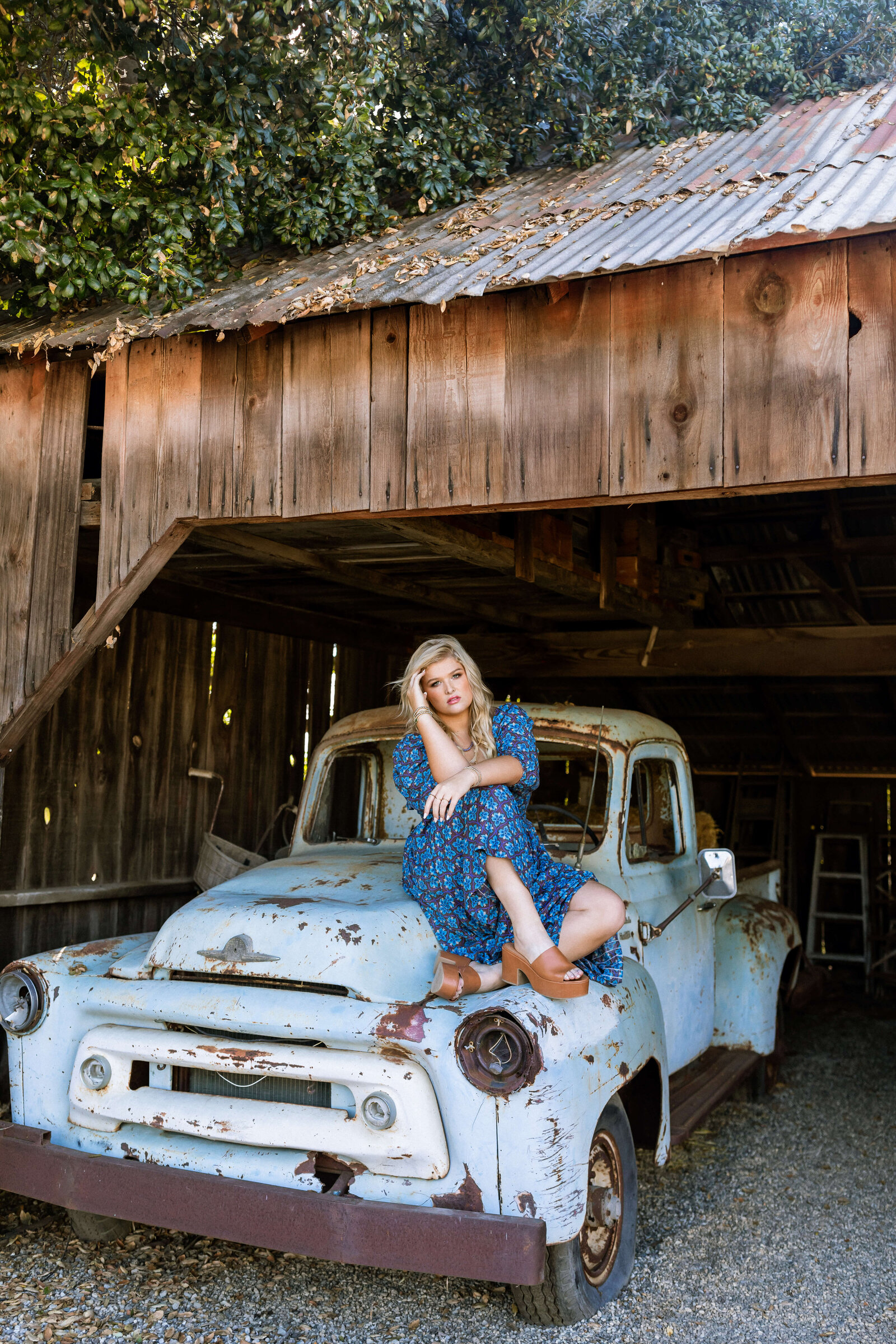 blonde girl wearing a blue and purple dress posing on an old rusted car in a barn