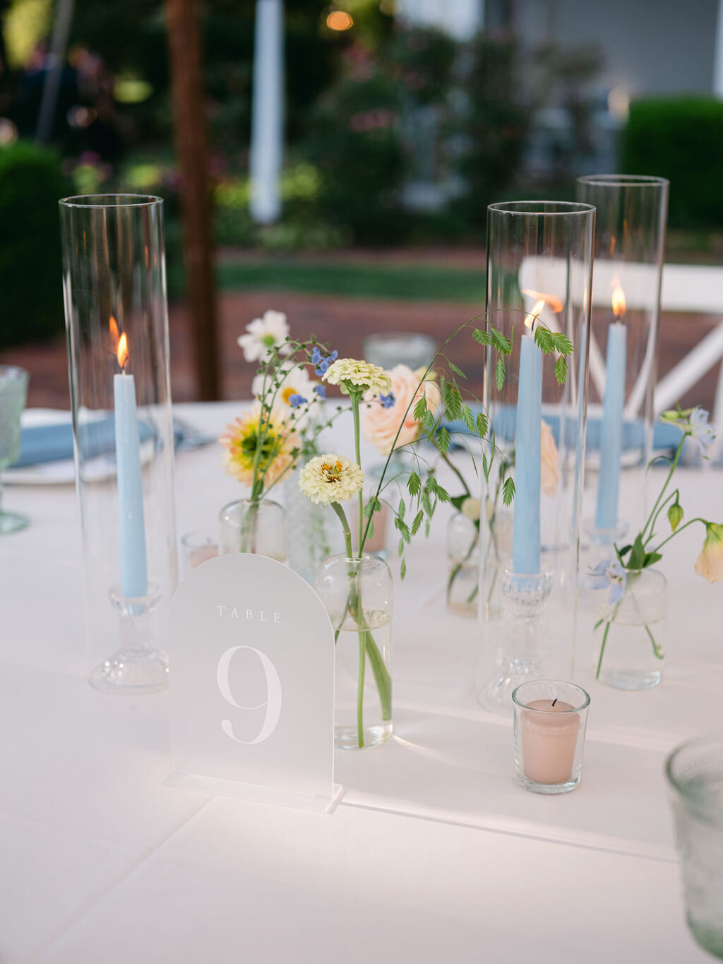 Guest table with accent florals such as white zinnias, white cosmos and lisianthus in bud vases alongside blue taper candles.
