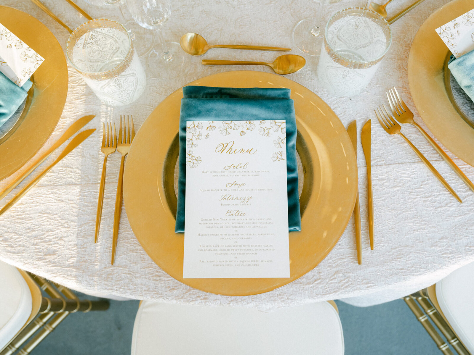A blue velvet napkin rests atop a gold charger that holds a menu. In the background, luxury glassware and candles create an elegant ambiance.