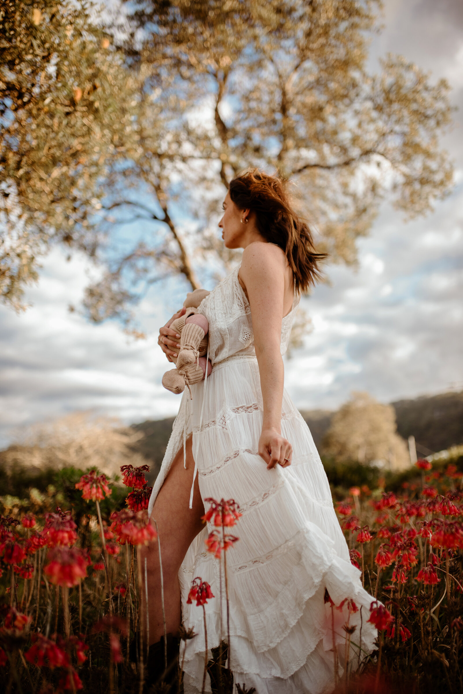 A woman stands in a field of red flowers, wearing a flowing white dress and holding a doll, with trees and a cloudy sky in the background.