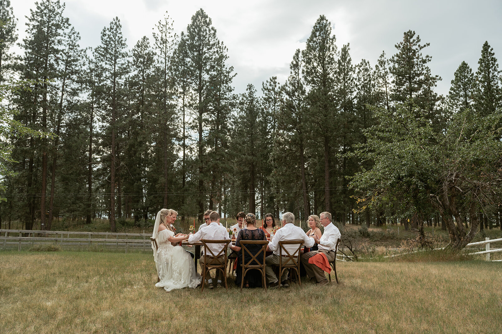 A small group of loved ones gathered around a wooden table outdoors, celebrating a newlywed couple’s intimate elopement surrounded by nature.