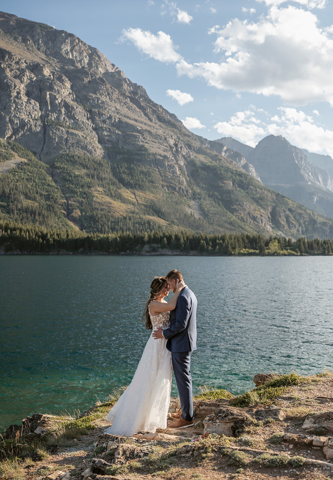 A couple embraces along the edge of a blue alpine lake surrounded by towering mountains and sunlight in Glacier National Park, captured by Sydney Breann Photography during their Montana elopement.