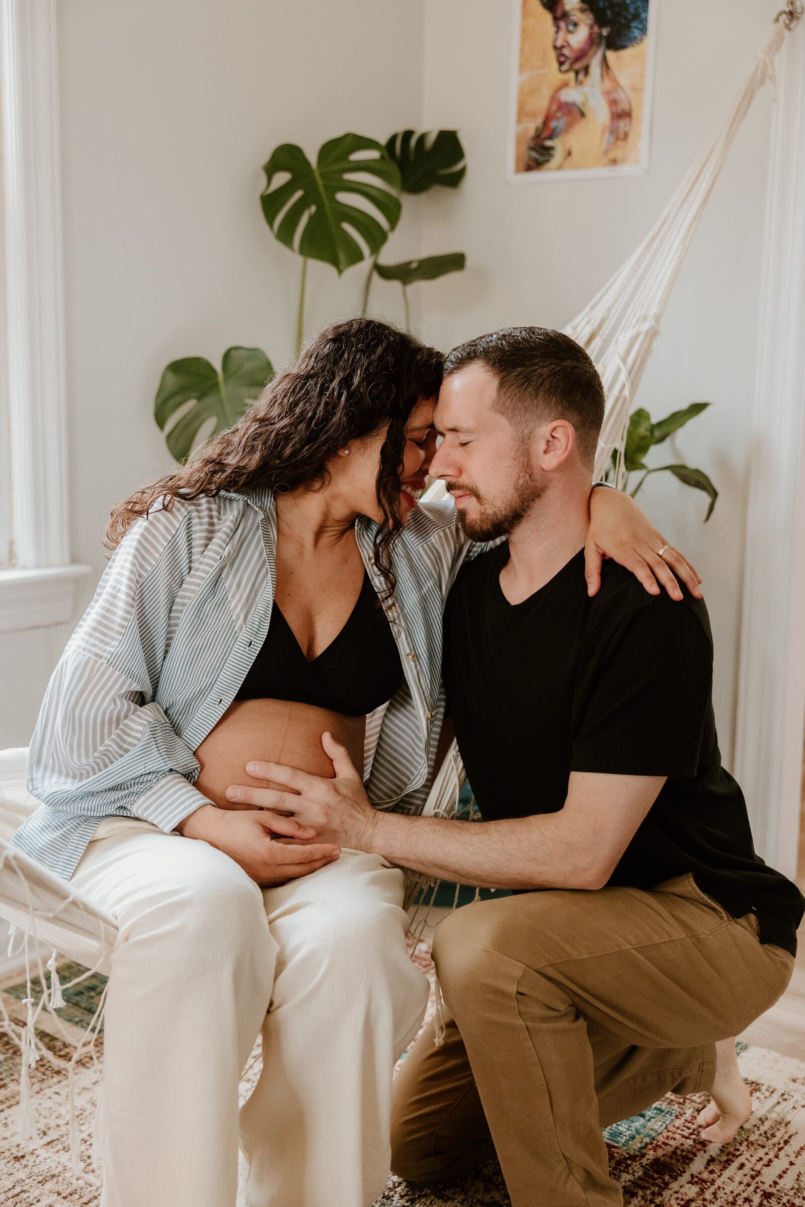 Expectant couple sharing a tender moment during an in-home maternity session; the woman sits in a hammock chair, cradling her baby bump, while the man kneels beside her with his hand on her belly. A monstera plant and artwork hang in the background, creating a cozy, intimate atmosphere.
