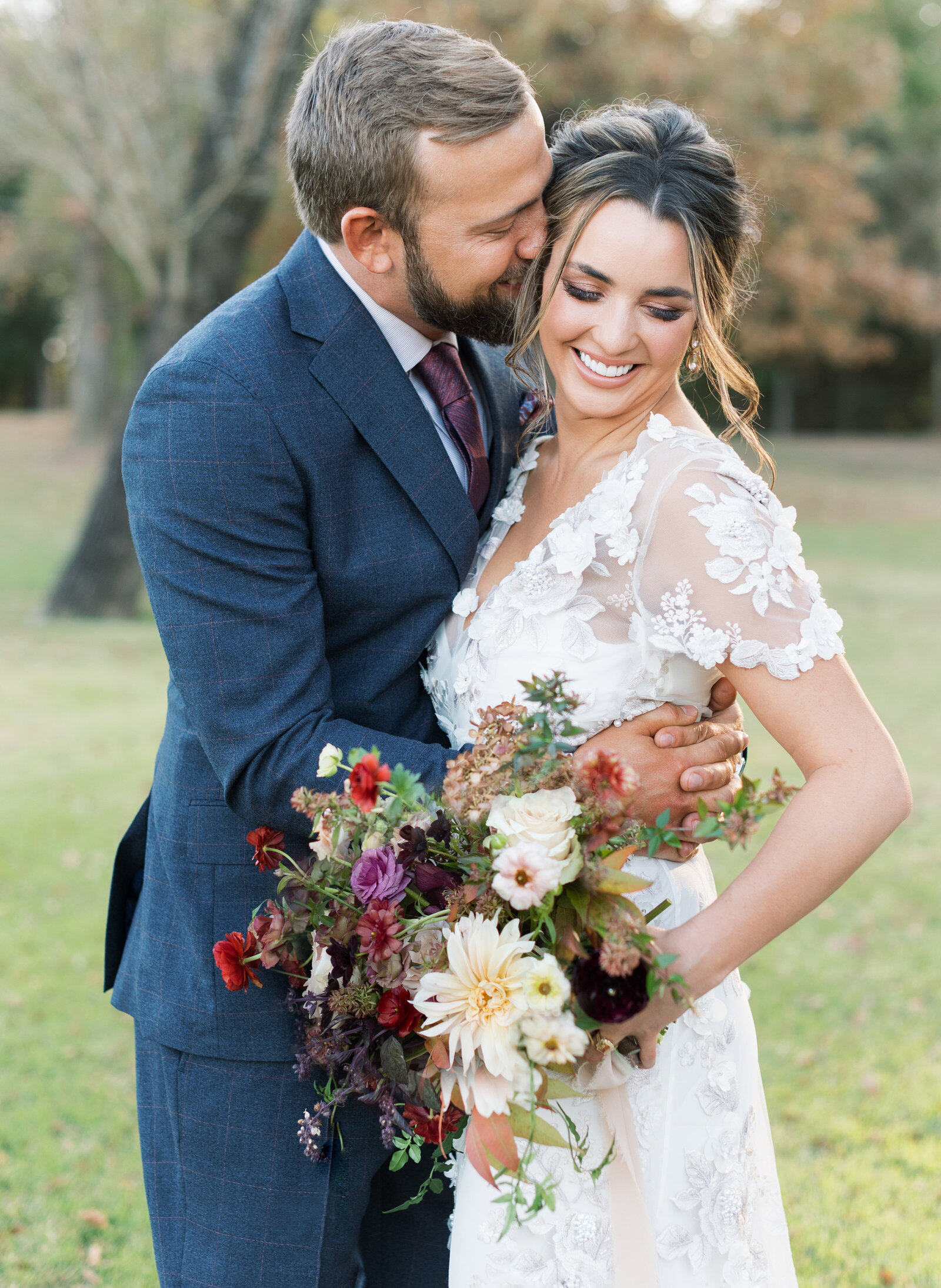 Portrait of a bride and groom in a white lace wedding gown and blue suit holding a bouquet outdoors atop the grass