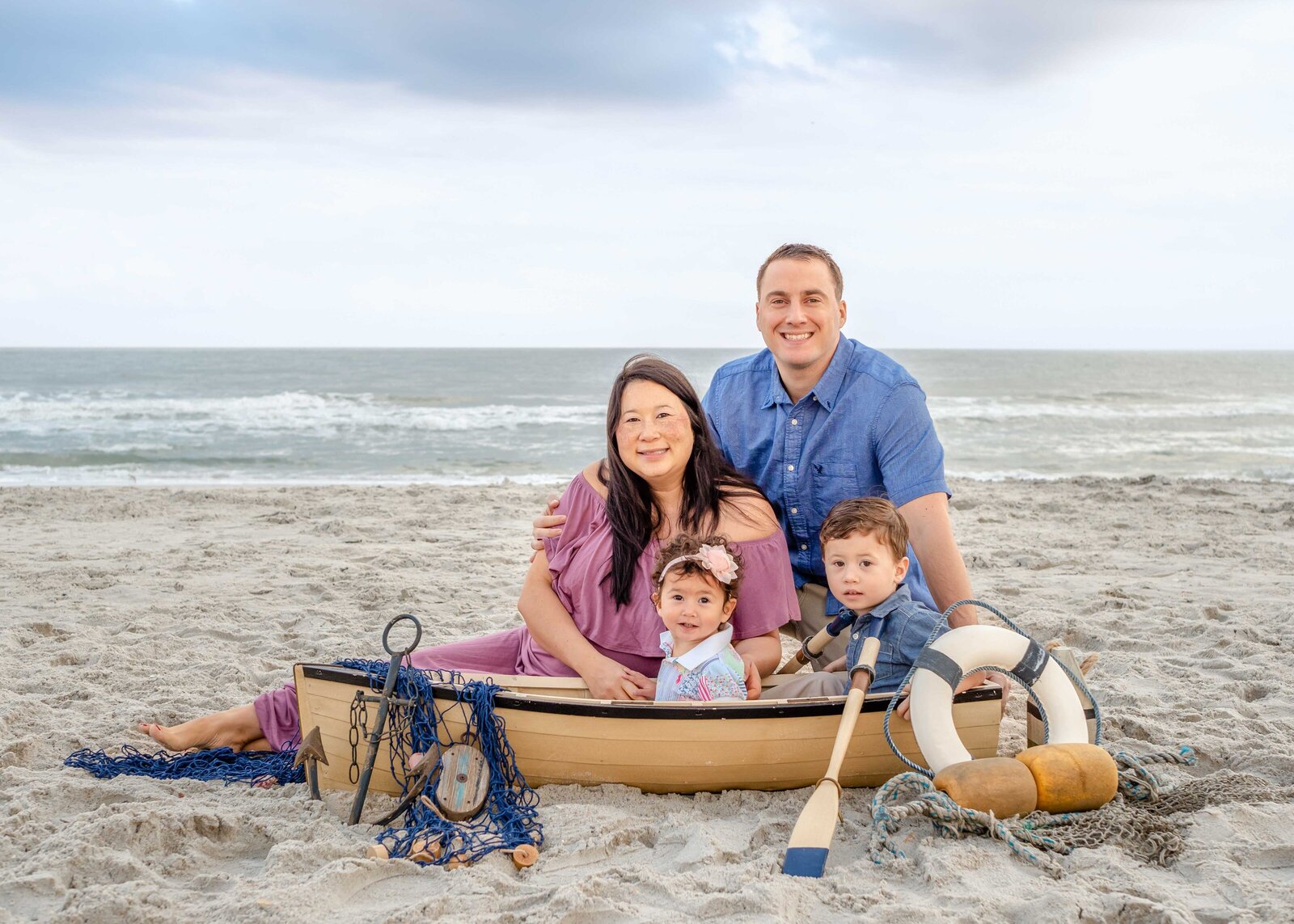 A family of four poses with a decorative boat prop on the sands of Carolina Beach. Dressed in pastel colors, they enjoy a whimsical and playful setting by the ocean. Perfect for professional family photography in NC, capturing creative beachside themes.