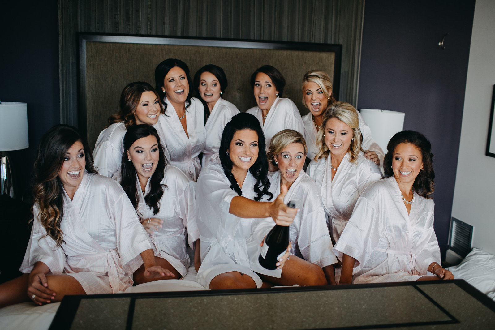 Bride and her bridesmaids bracing for a uncorking of a Champagne bottle in a hotel room.