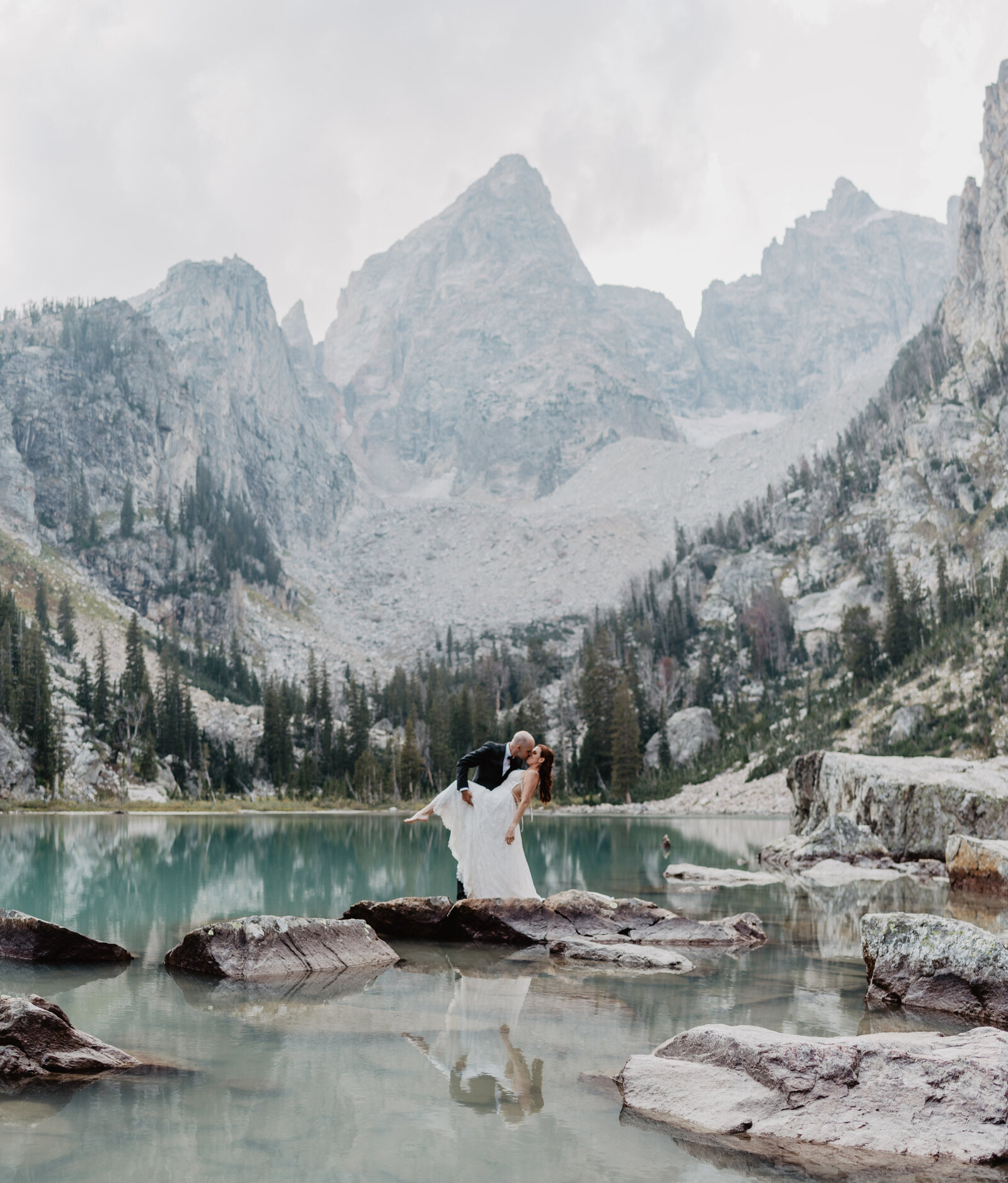Delta Lake Elopement with bride and groom kissing passionately in the middle of the lake with the grand tetons perfectly framing the bright blue lake with a  small woods at the base of the mountain