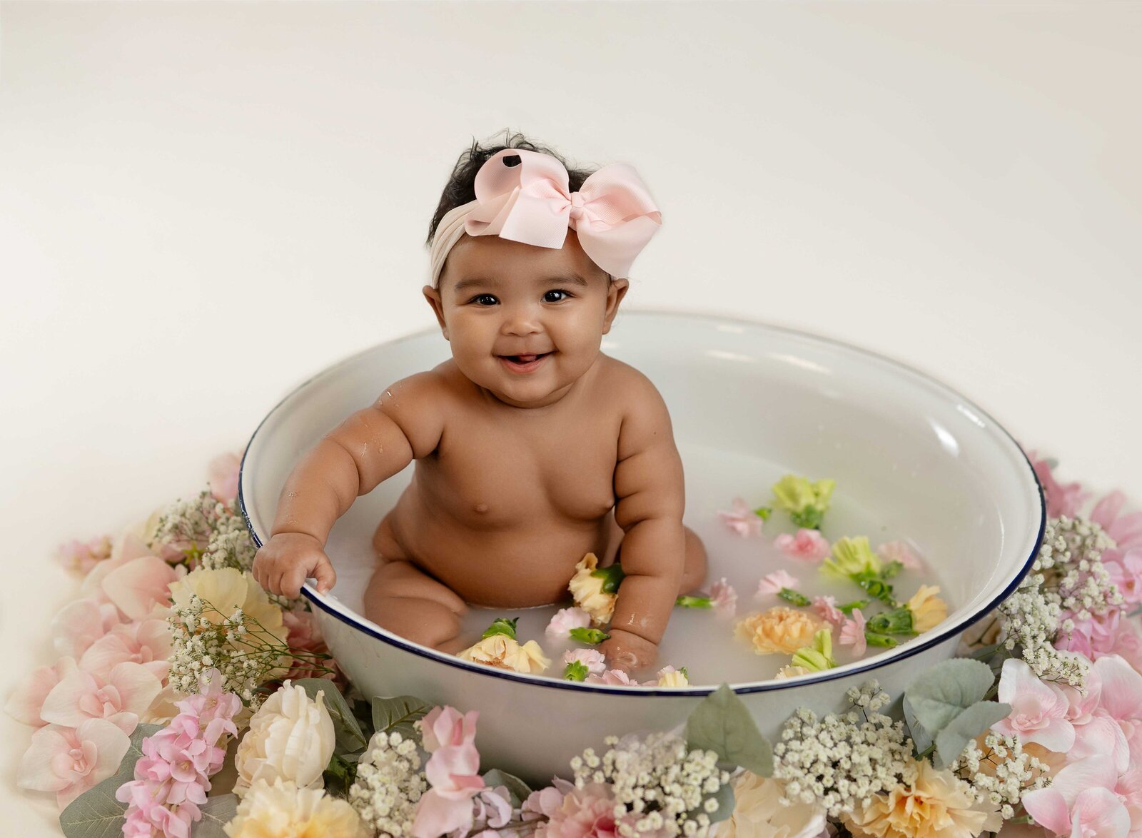 Baby girl sitting in a basin filled with a milk bath and flowers and smiling.