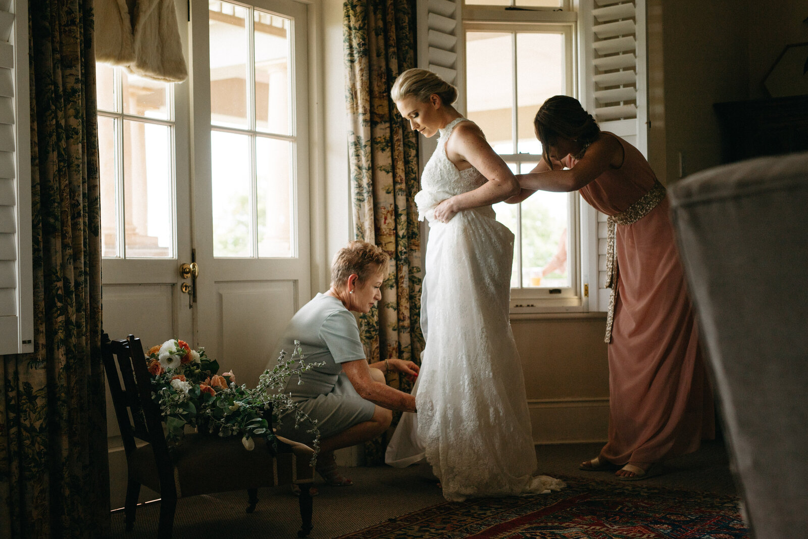 A bride is helped into her dress by her mother and bridesmaid,  they are lit softly by the window light in a tender moment