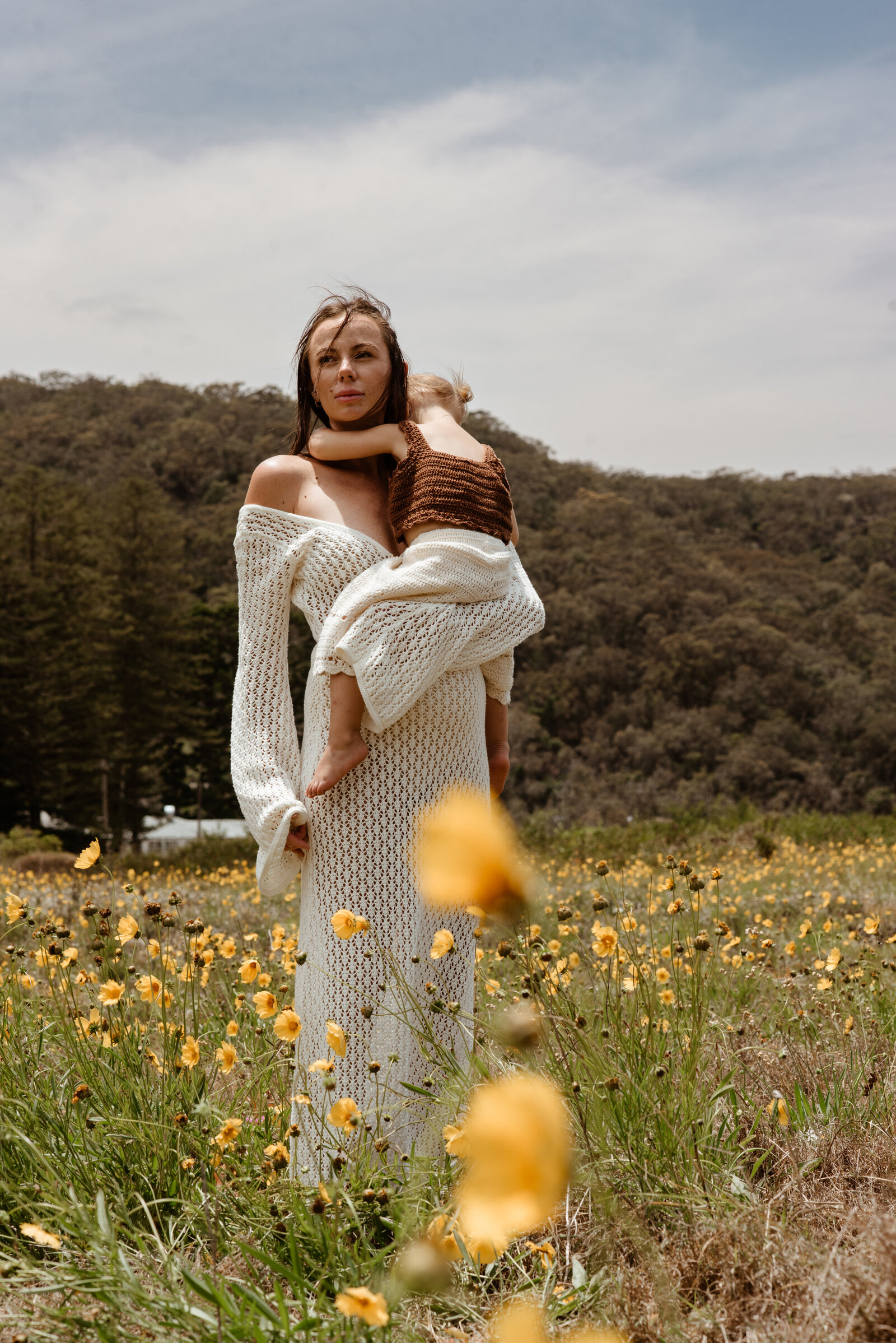 A woman in a white crochet dress stands in a field of yellow flowers, holding a child who is wearing a brown outfit, with a forested hill in the background.