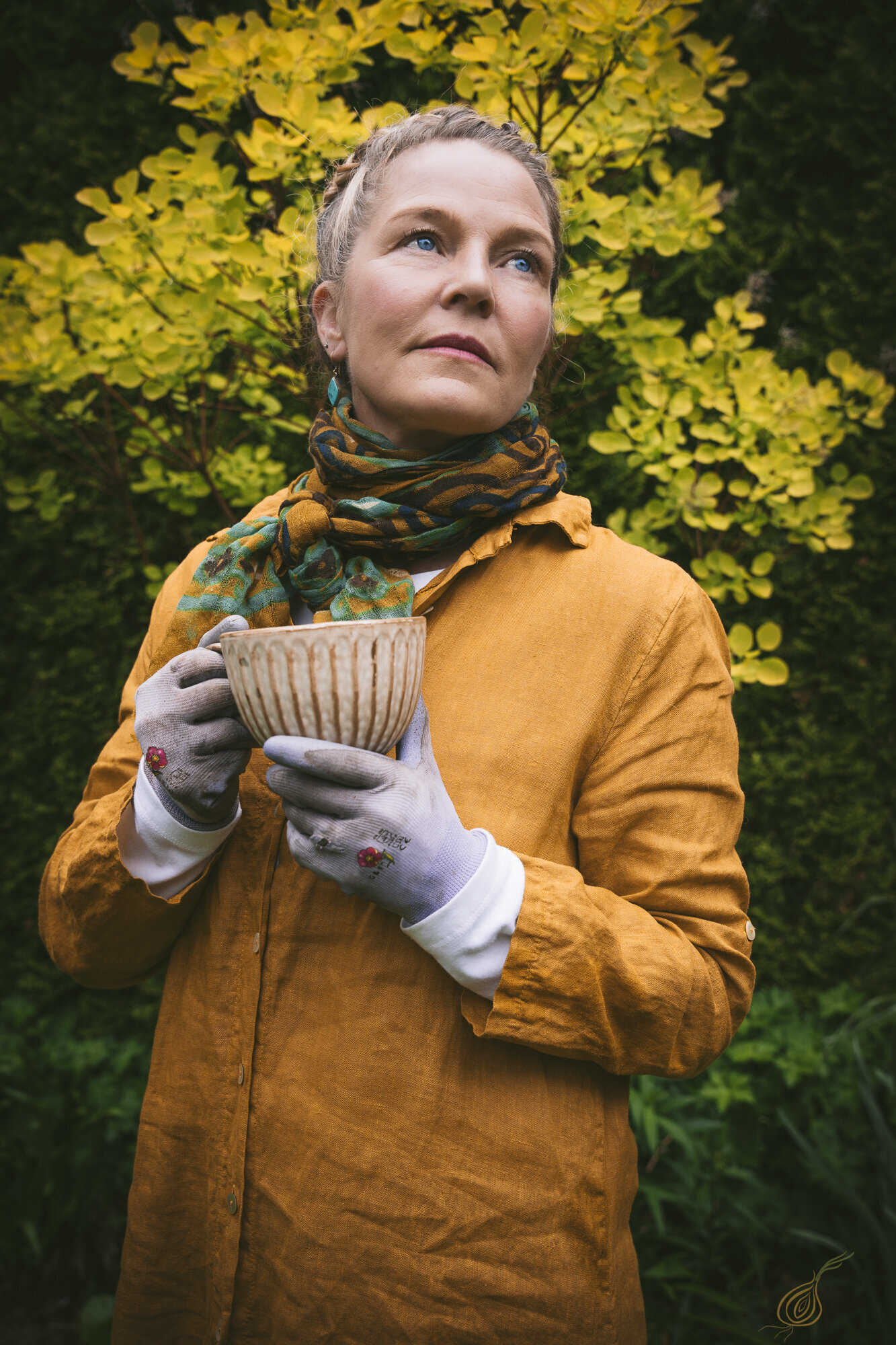 A woman gracefully holds a rustic cup that contains tea as she stands behind an assortment of colorful flowers.