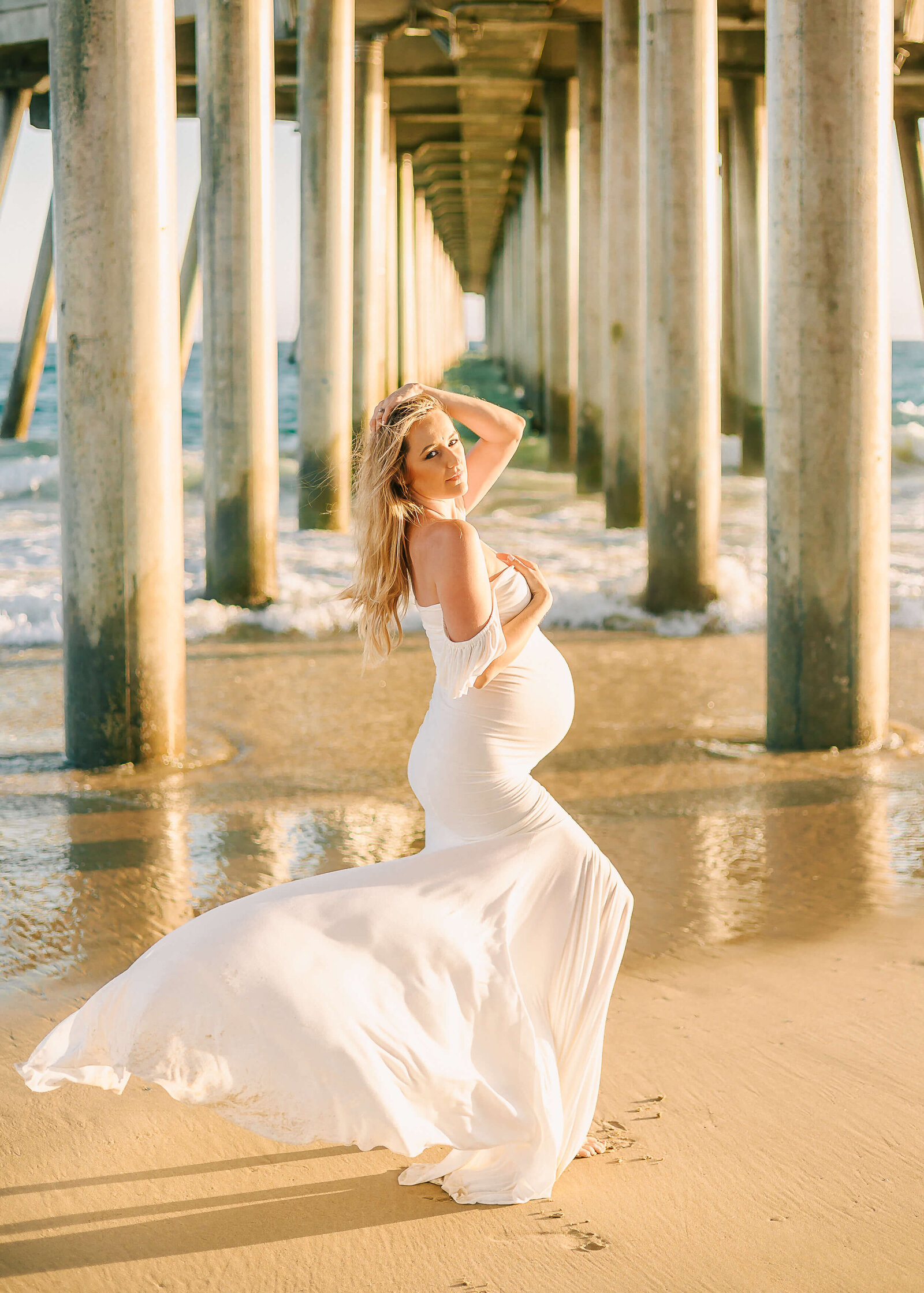 Mama wearing white fitted dress during maternity session at Huntington Beach Pier with the wind blowing her hair and the dress back.