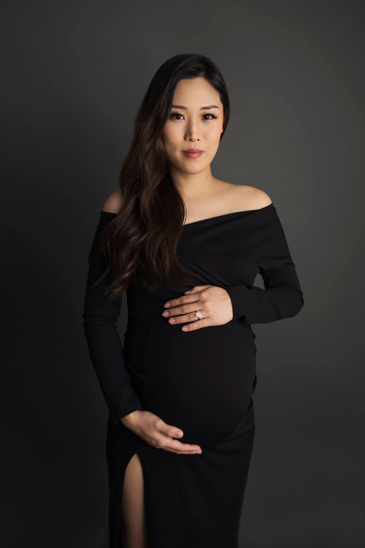 A pregnant woman stands against a dark background, wearing an off-the-shoulder, long-sleeved black gown with a thigh-high slit. Her long, dark hair falls in loose waves over one shoulder. She cradles her baby bump with both hands, displaying a calm and confident expression. The dark tones of the portrait highlight the elegance and simplicity of the moment.