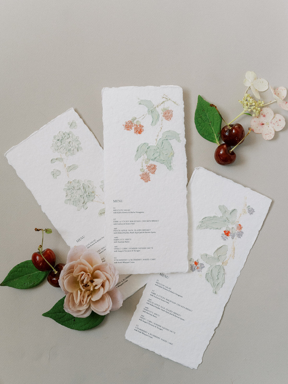 Flat lay of custom hand-painted menus with blush garden rose and cherries.