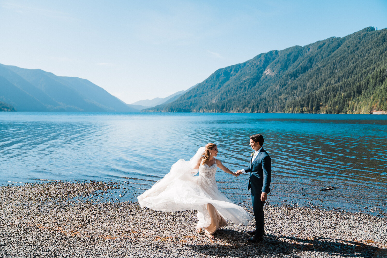 A groom twirls his bride along the shores of Lake Crescent in Olympic National Park on a bright sunny day photographed by PNW elopement photographer Amy Galbraith