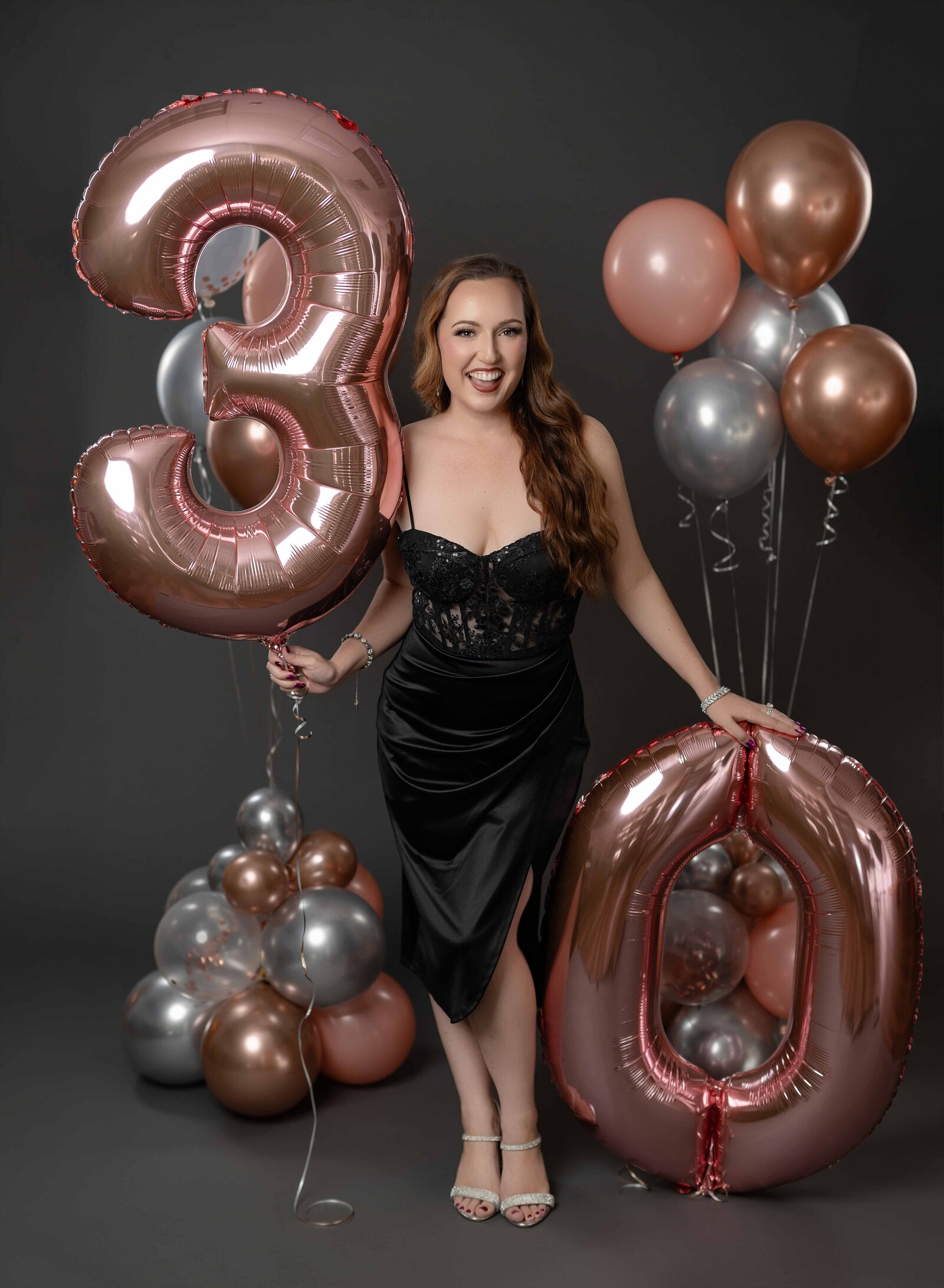 Women in black dress, posing for 30th birthday photo and holding #30 rose gold balloons. Photo taken at Atlanta Birthday photo studio.