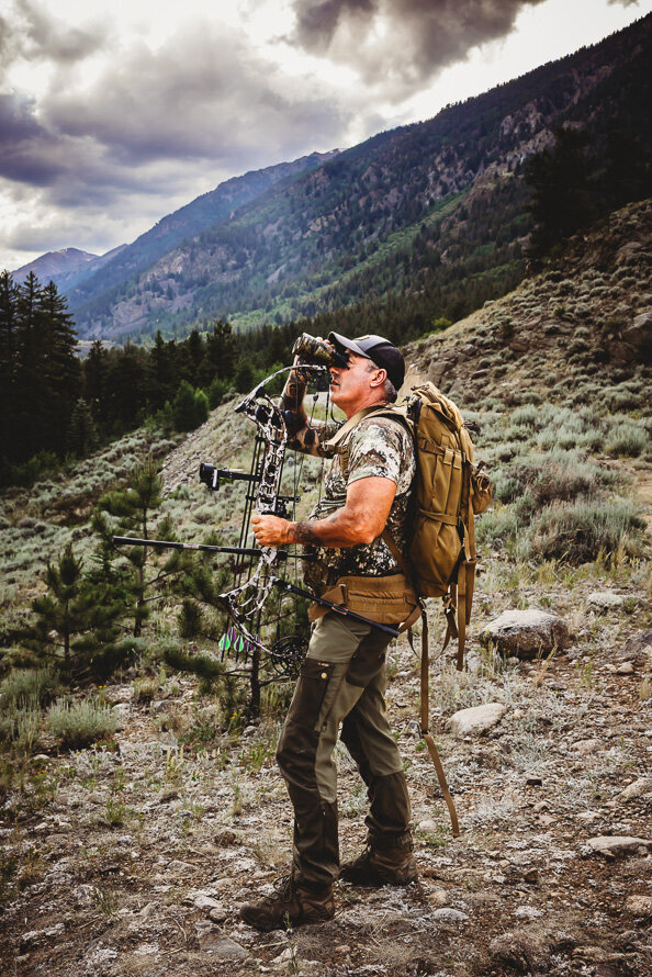 Bow hunter in camouflage looks through is binoculars with mountains behind him.