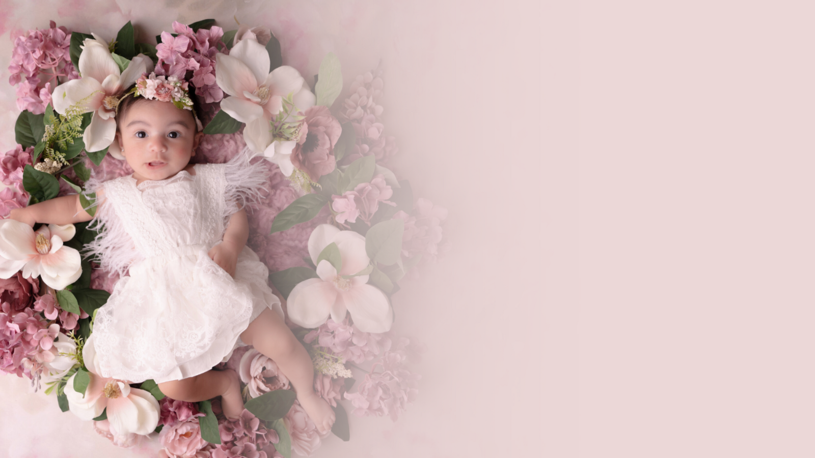 A baby in a white, lace dress lies among soft pink and white flowers with green leaves. The background fades to a light pink, creating a serene and gentle ambience.