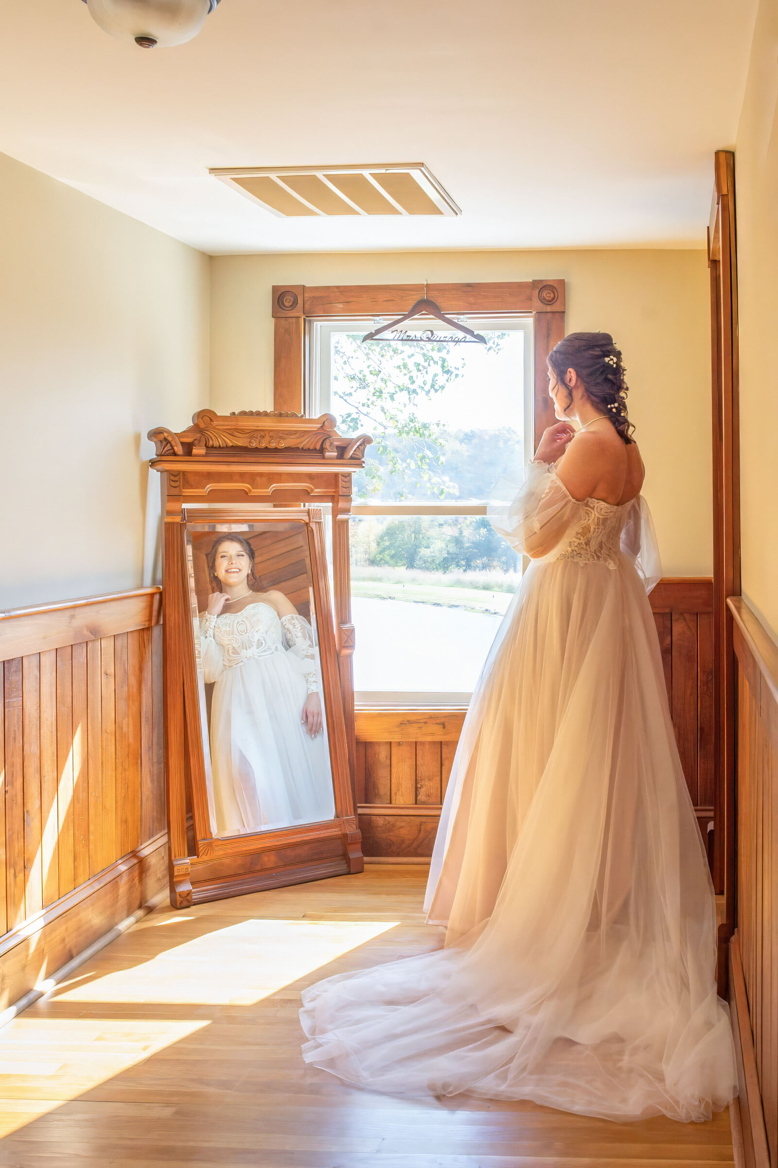 A bride in a flowing gown admires her reflection in a vintage mirror surrounded by rustic wooden details, captured in a bridal suite during a Wilmington, NC wedding, blending elegance and authenticity.