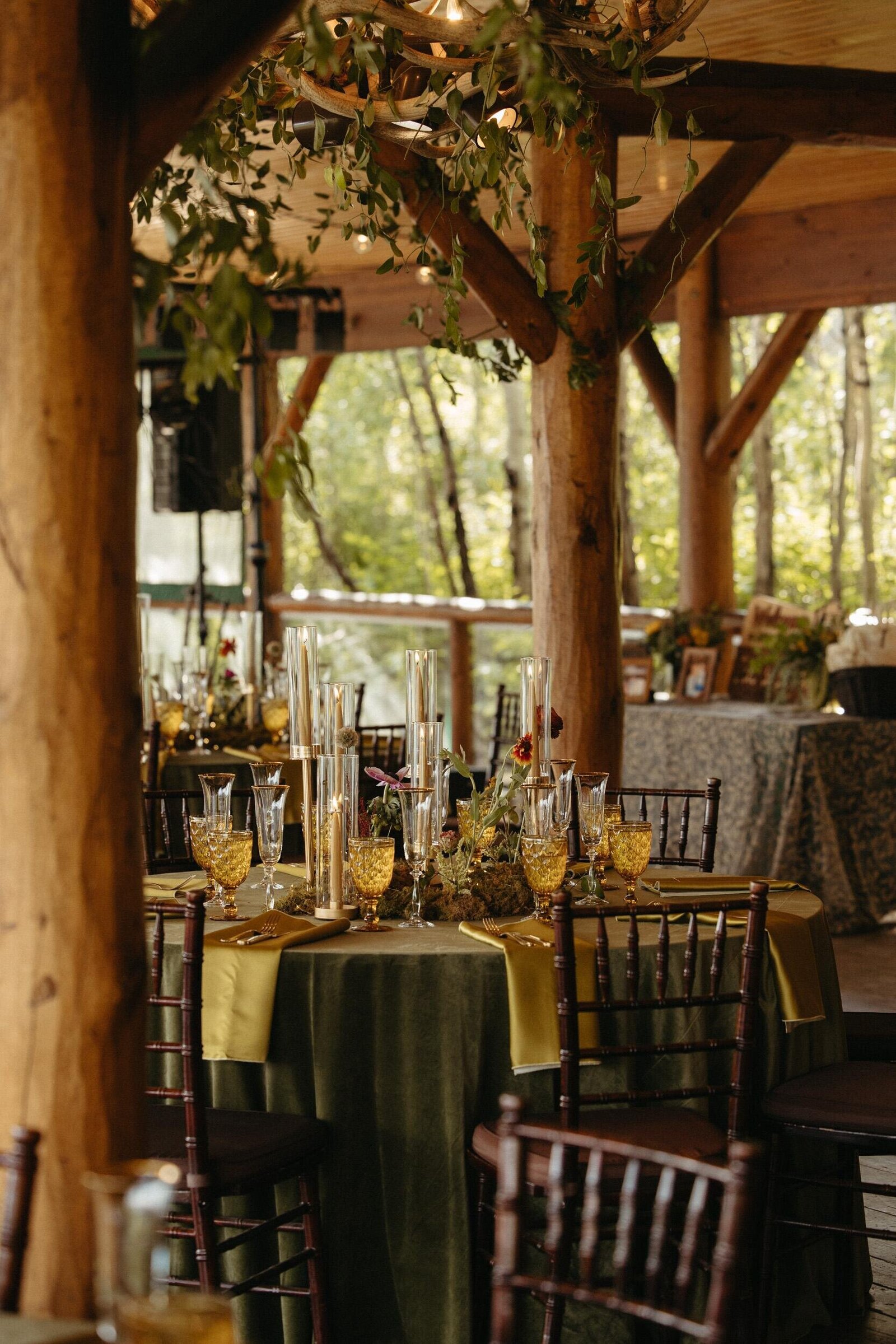 Woodsy wedding venue with green velvet table linens, gold napkins, mossy centerpieces and gold candles in glass candelabras