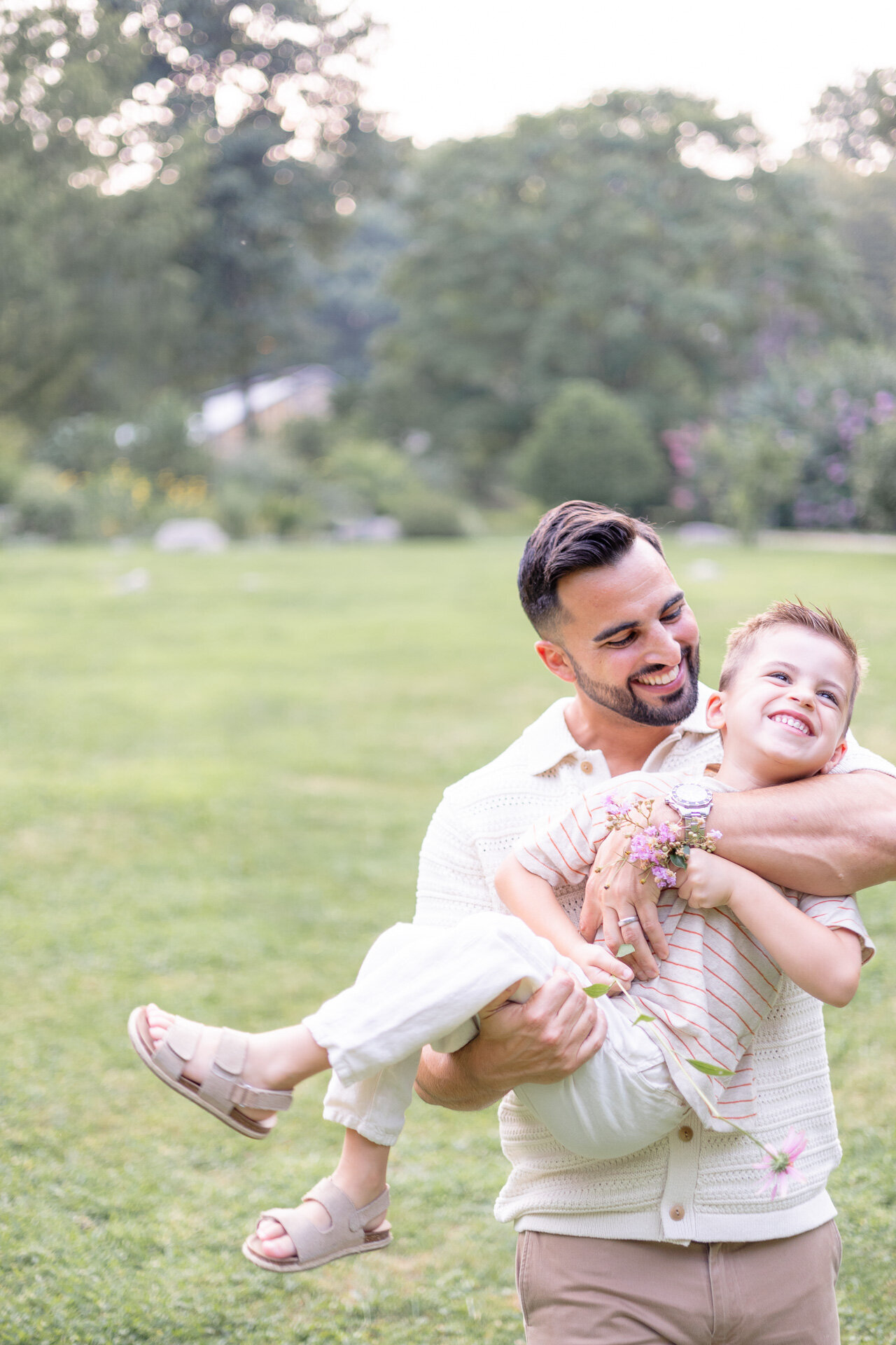 A joyful father twirls his smiling son in an open grassy field during a summer family photography session in Fairfield County, CT. The moment is full of laughter and connection, with wildflowers in the background and a flower bracelet on the child’s wrist.