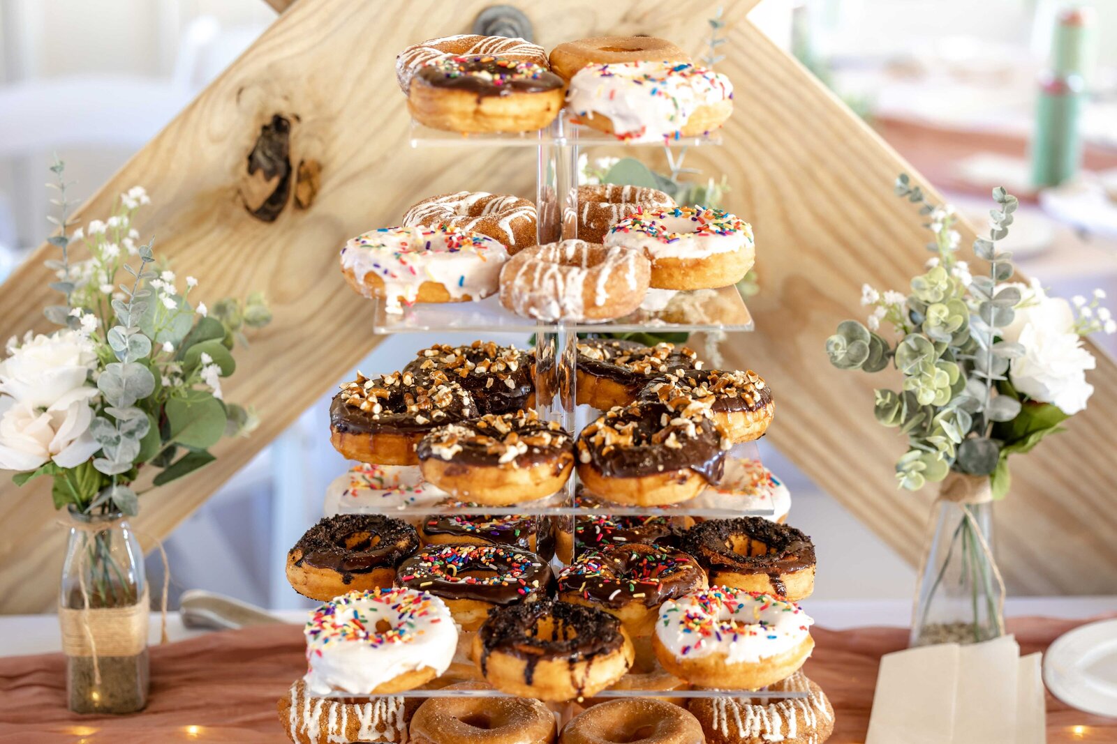A tiered wedding donut tower decorated with colorful sprinkles and rustic wooden accents, surrounded by floral arrangements, captured at a Wrightsville Beach, NC wedding, blending fun and rustic charm.