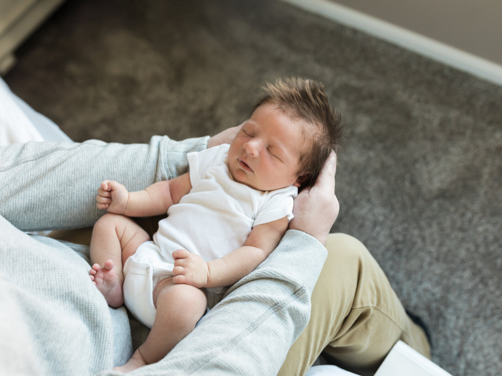 newborn baby boy in white onesie laying in dads lap for in home lifestyle session cleveland newborn photographer