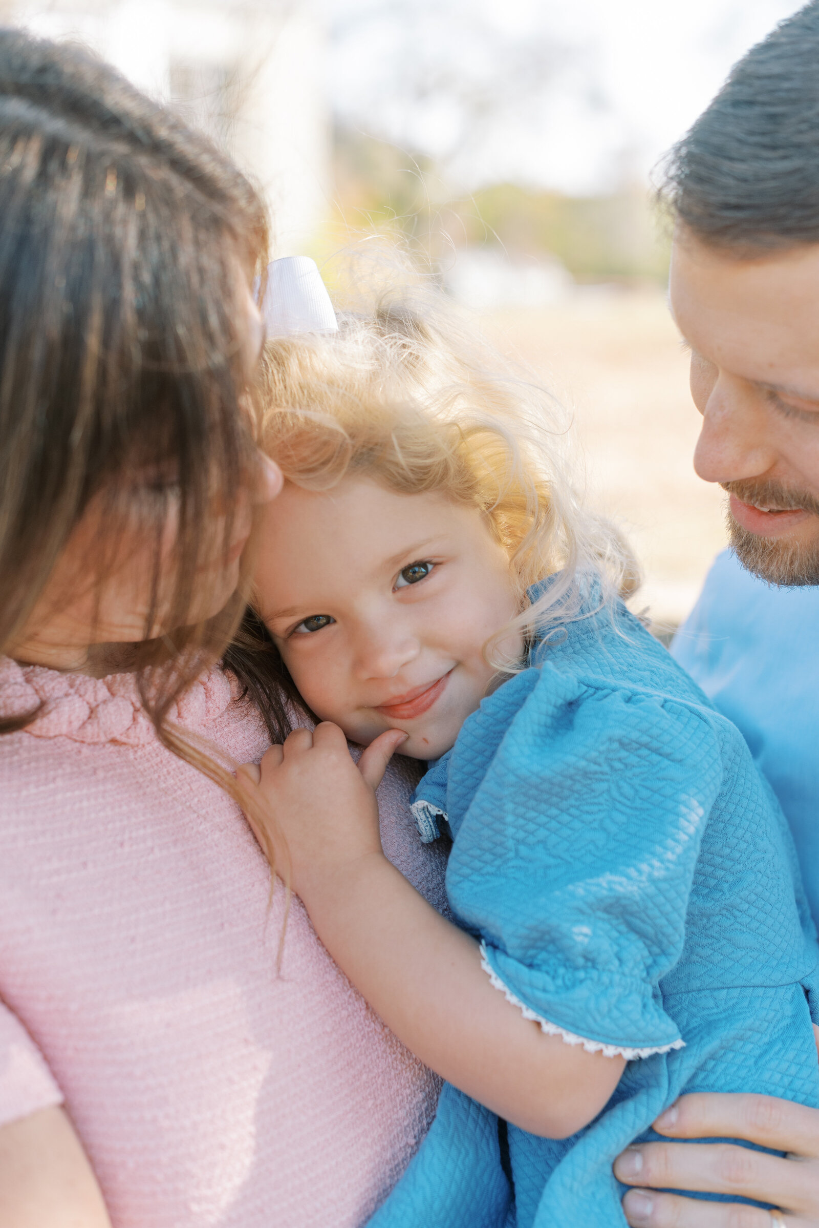 Photo of a family, close up of a baby girl in a blue dress smiling while her parents look on by Richmond family photographer