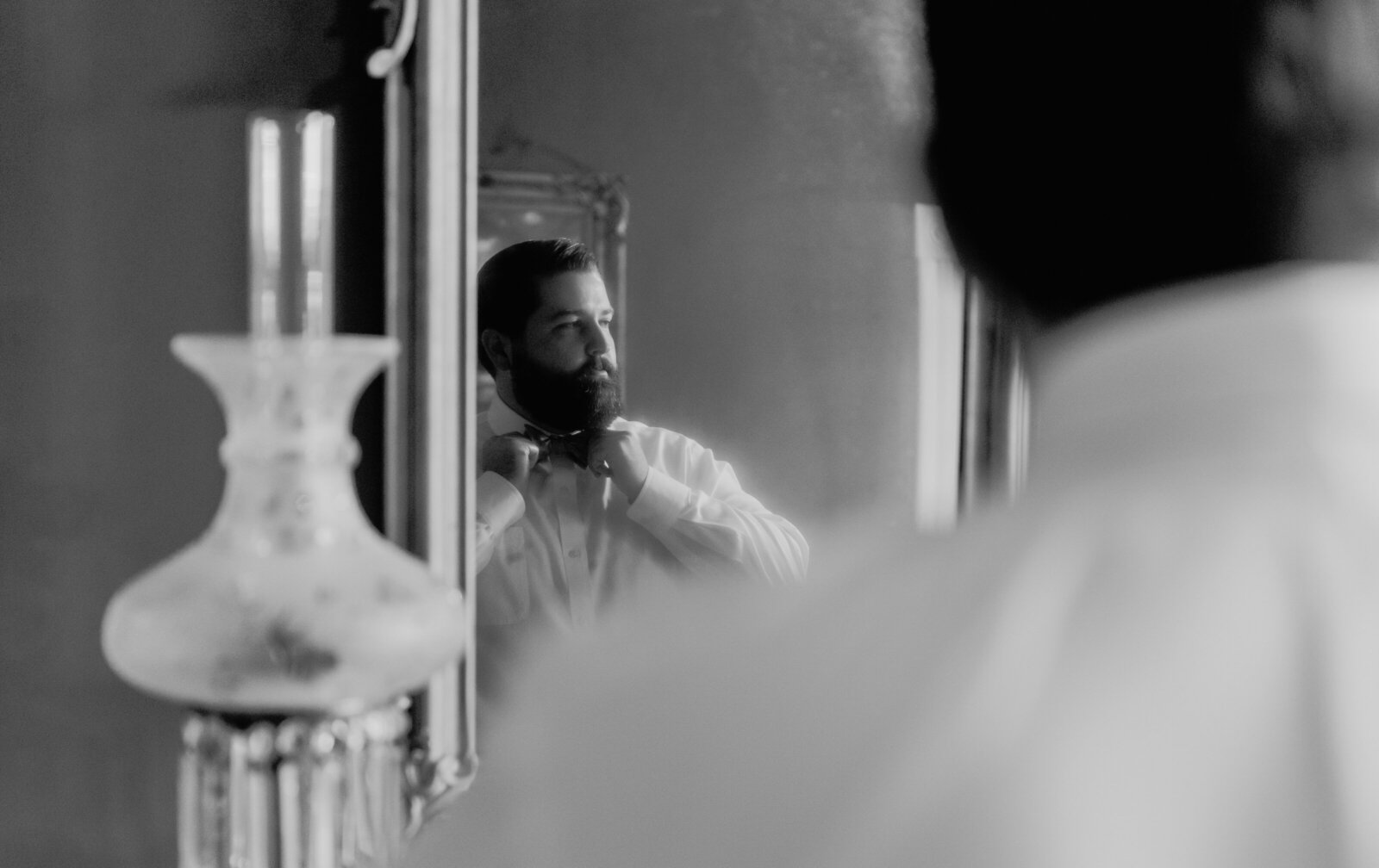 A groom is reflected in a mirror while fixing his bowtie