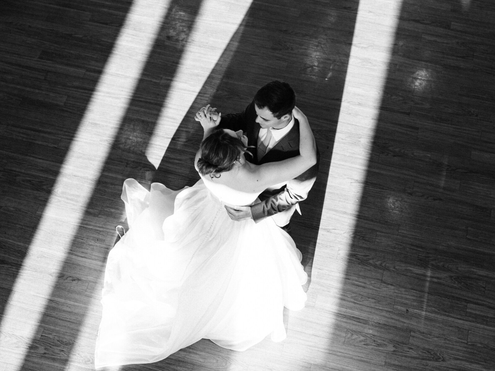 A bride and groom dance together on a wooden floor, bathed in sunlight streaming through windows, casting dramatic shadows around them. The brides dress fans out elegantly in the image by an Austin wedding photographer.