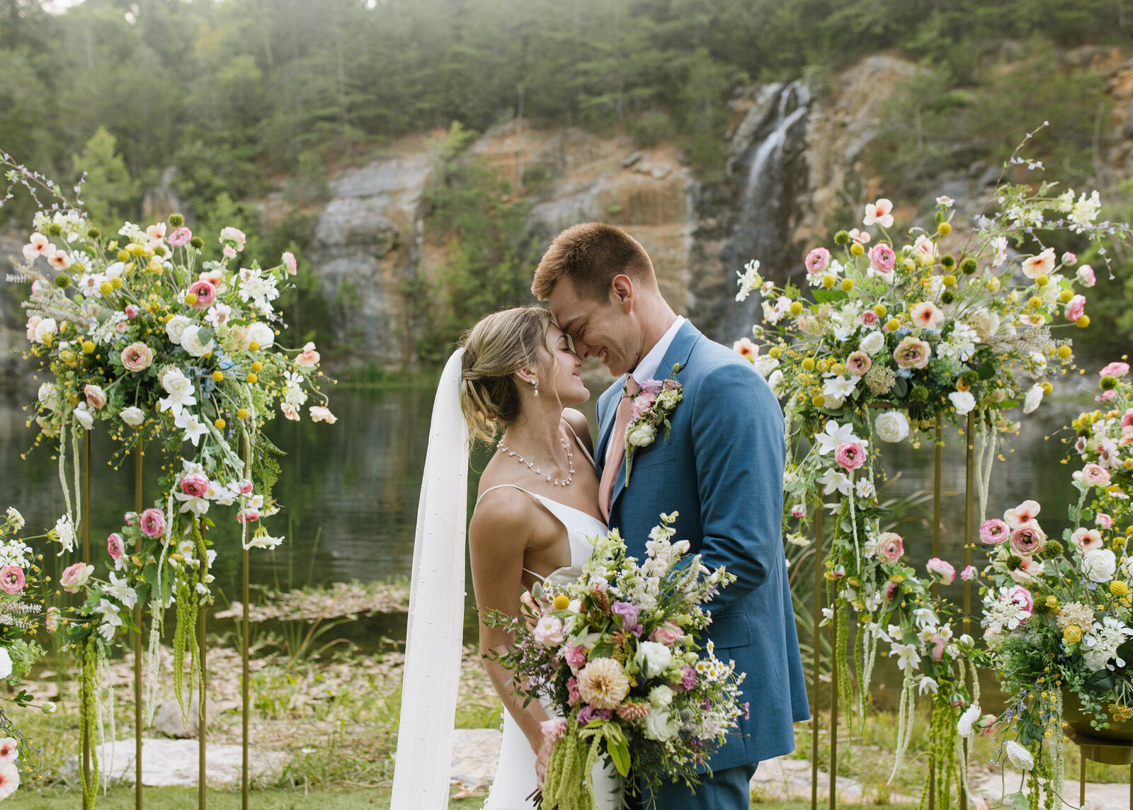A candid moment between the bride and groom during their vibrant summer wedding in East Tennessee. Surrounded by colorful wildflower-inspired florals and a stunning quarry waterfall backdrop, this true-to-color image captures the heart of documentary-style wedding photography.