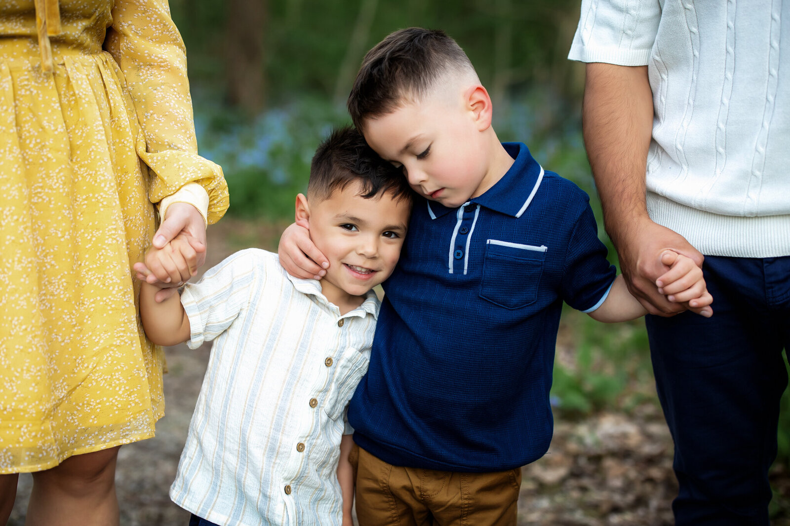 Two young boys stand close together outdoors, holding hands with adults on each side. The older boy in blue hugs the younger boy in white, who smiles at the camera—capturing a candid moment perfect for outdoor portraits.