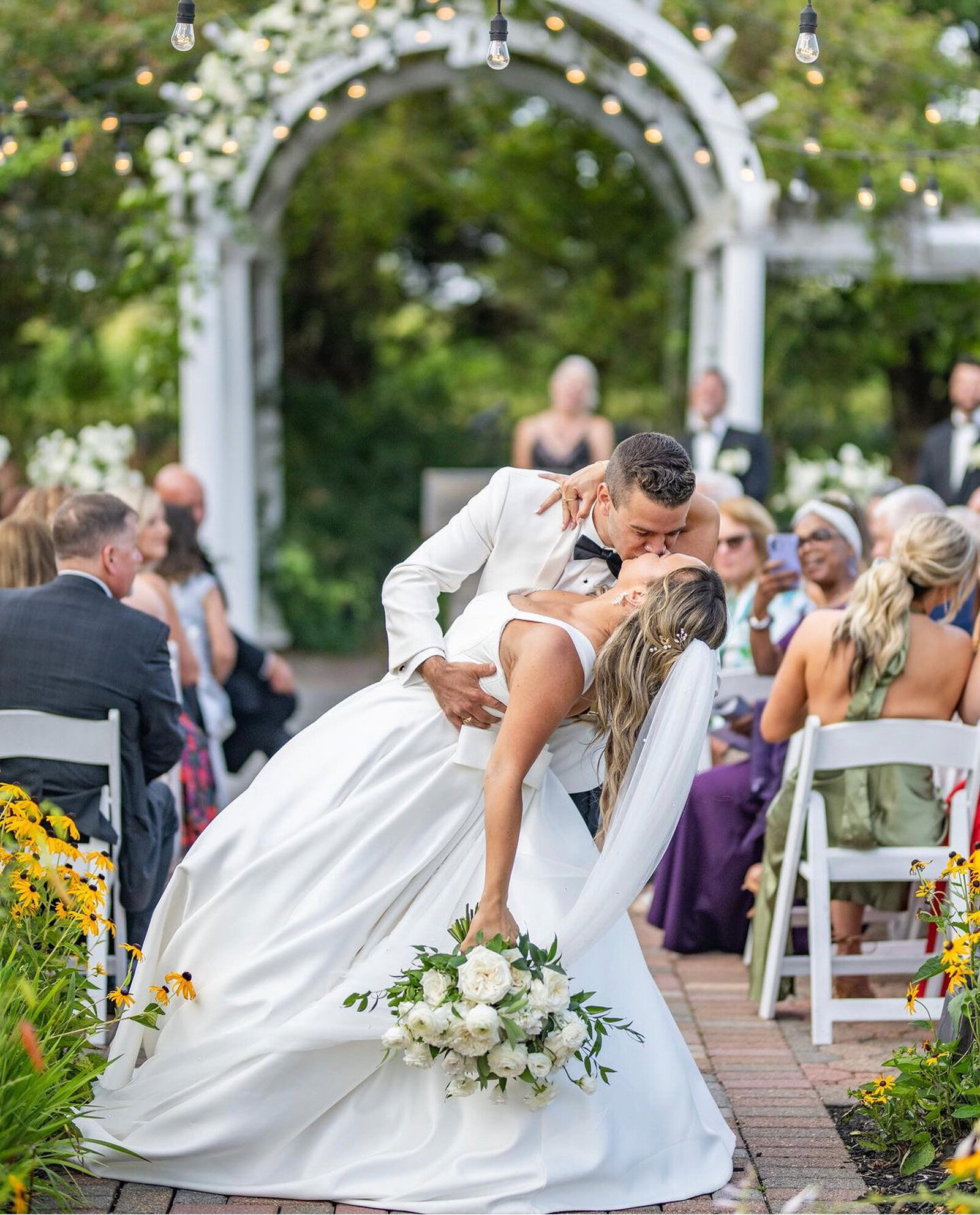 groom in a white tuxedo romantically dips his bride, who is wearing a flowing white wedding gown, as they share a kiss at an outdoor ceremony. Guests seated on white chairs and floral decorations are visible in the background