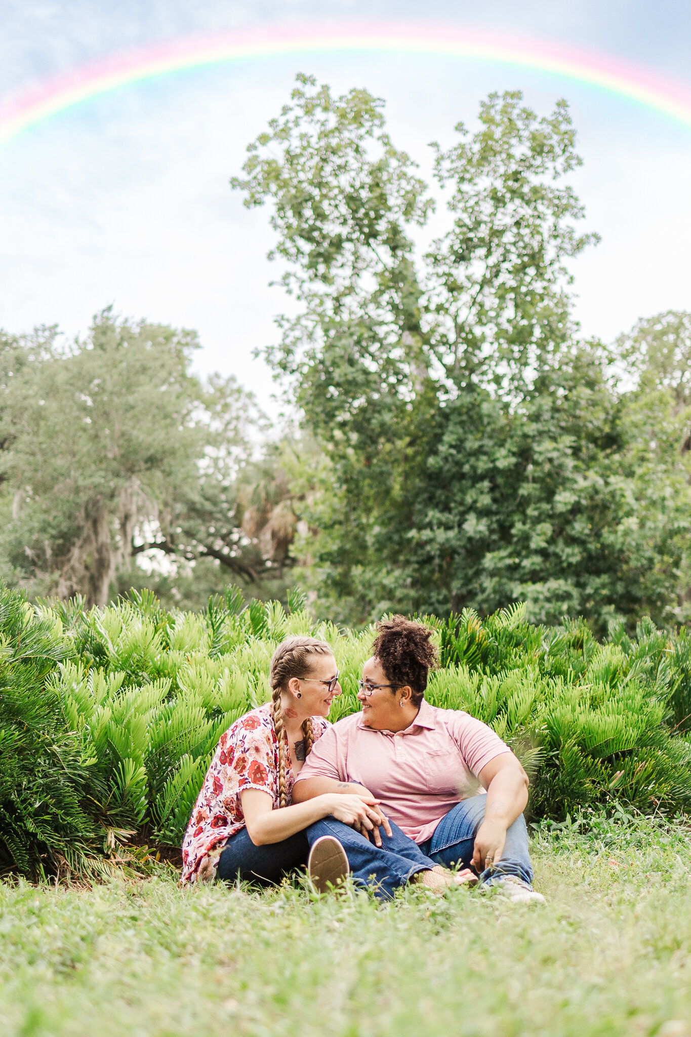 An interracial lesbian couple sitting on the grass with a rainbow