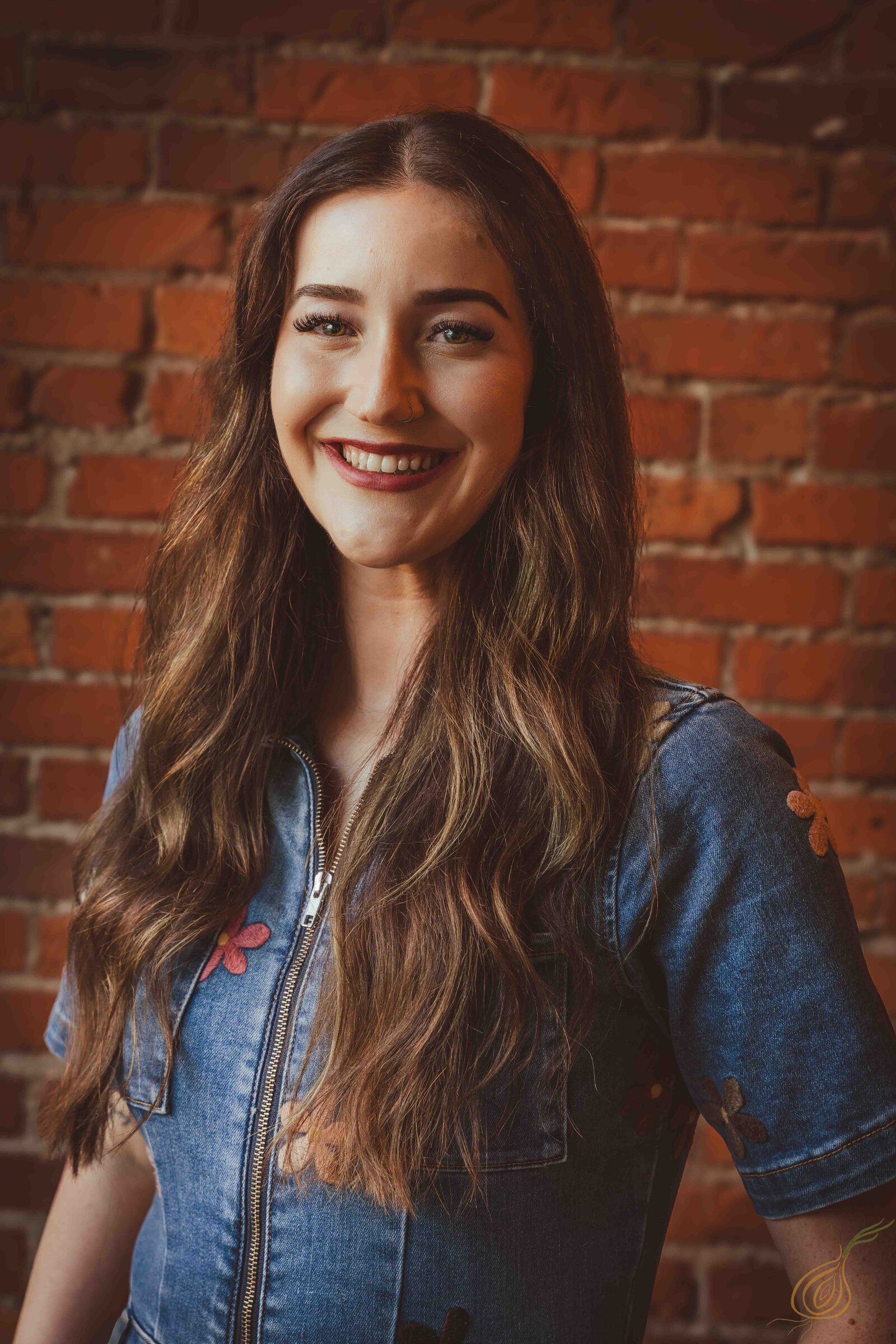 A woman with long hair wearing a denim shirt, standing confidently against a brick wall background.