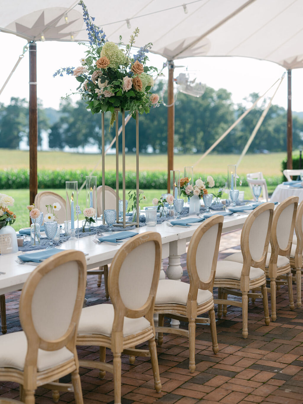 Long head table with large arrangement on a gold stand with florals including green hydrangea, blue delphinium, toffee roses, blush garden roses, and green hanging amaranthus suspended over a tablescape of blue taper candles and floral accents in bud vases.