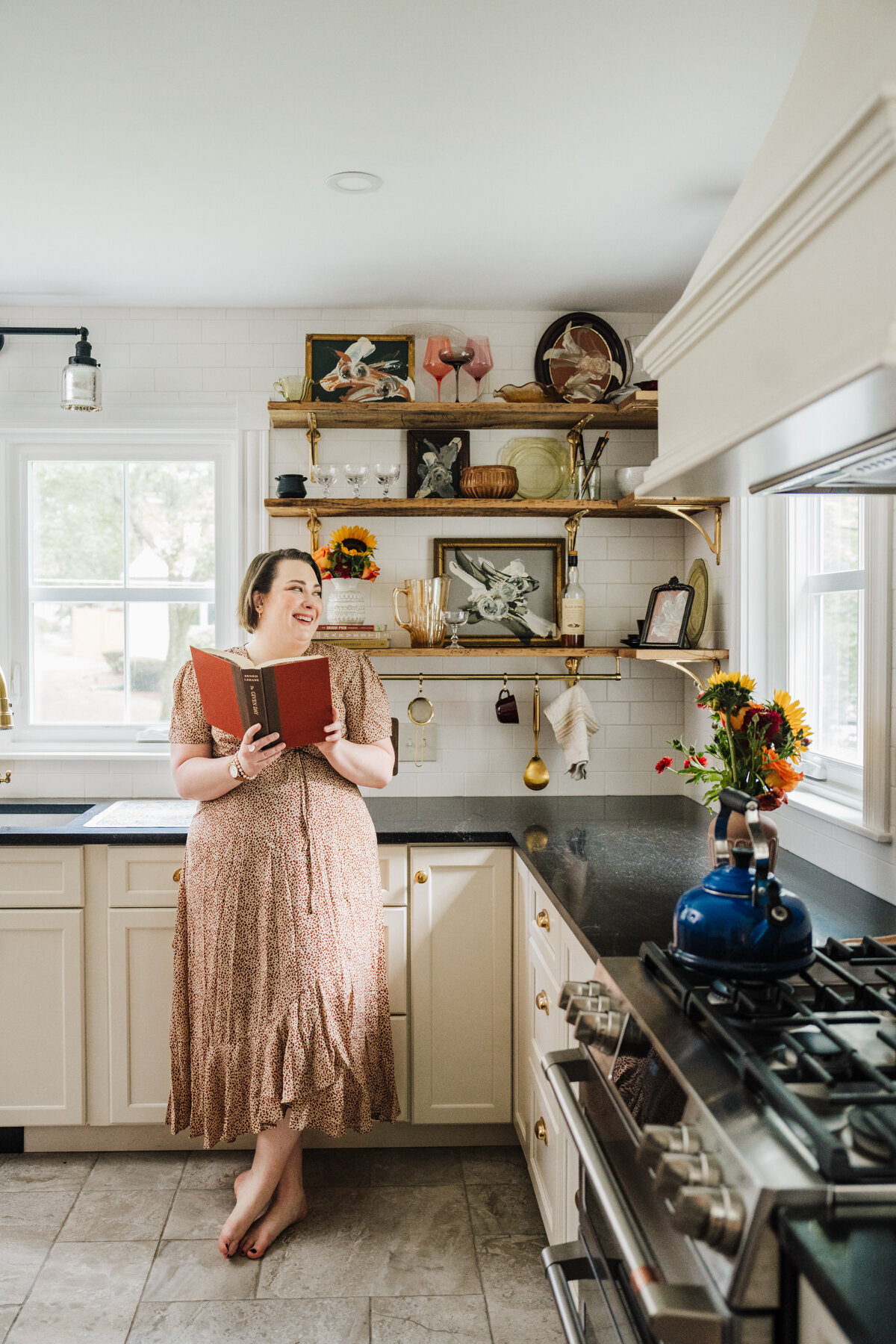 woman in dress reads book and leans against kitchen counter