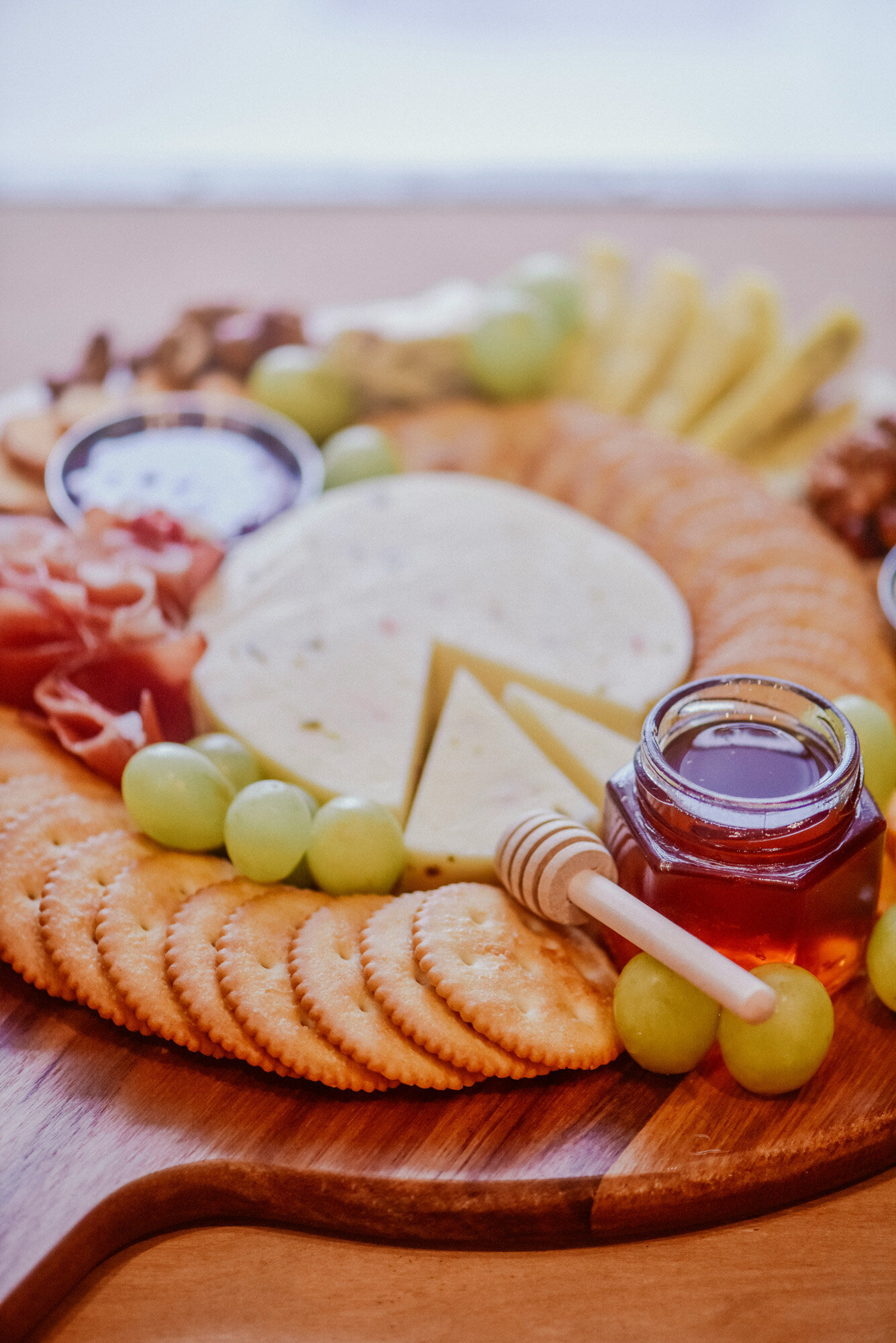 charcuterie board featuring sliced cheese, crackers, grapes, prosciutto, small jars of honey and fruit preserves