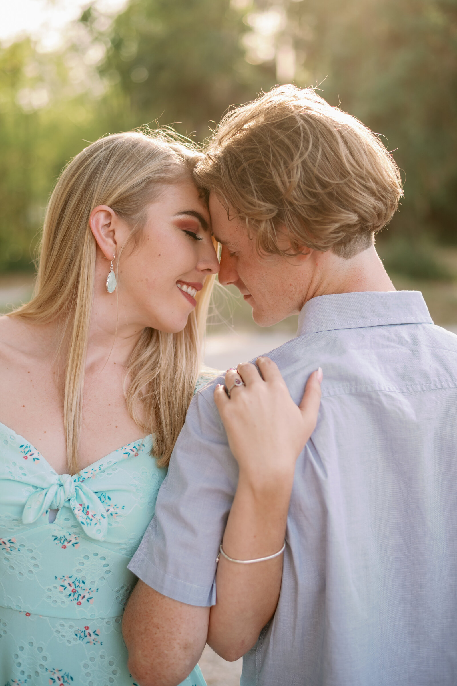 engaged couple touching foreheads together, his back is toward the camera while she is turned toward the camera. Both are smiling with eyes closed