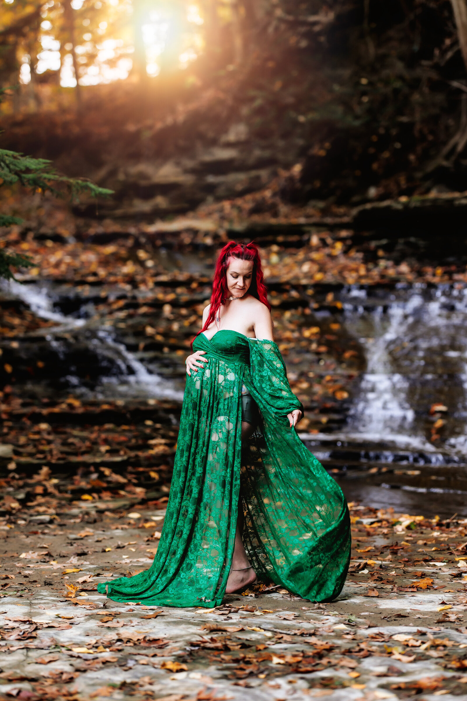 Pregnant mother wearing a long green lace dress, poses amongst a waterfall and beautiful fall foliage