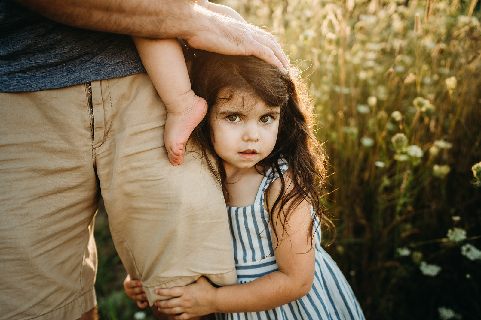little girl looks straight into camera while hugging dads leg