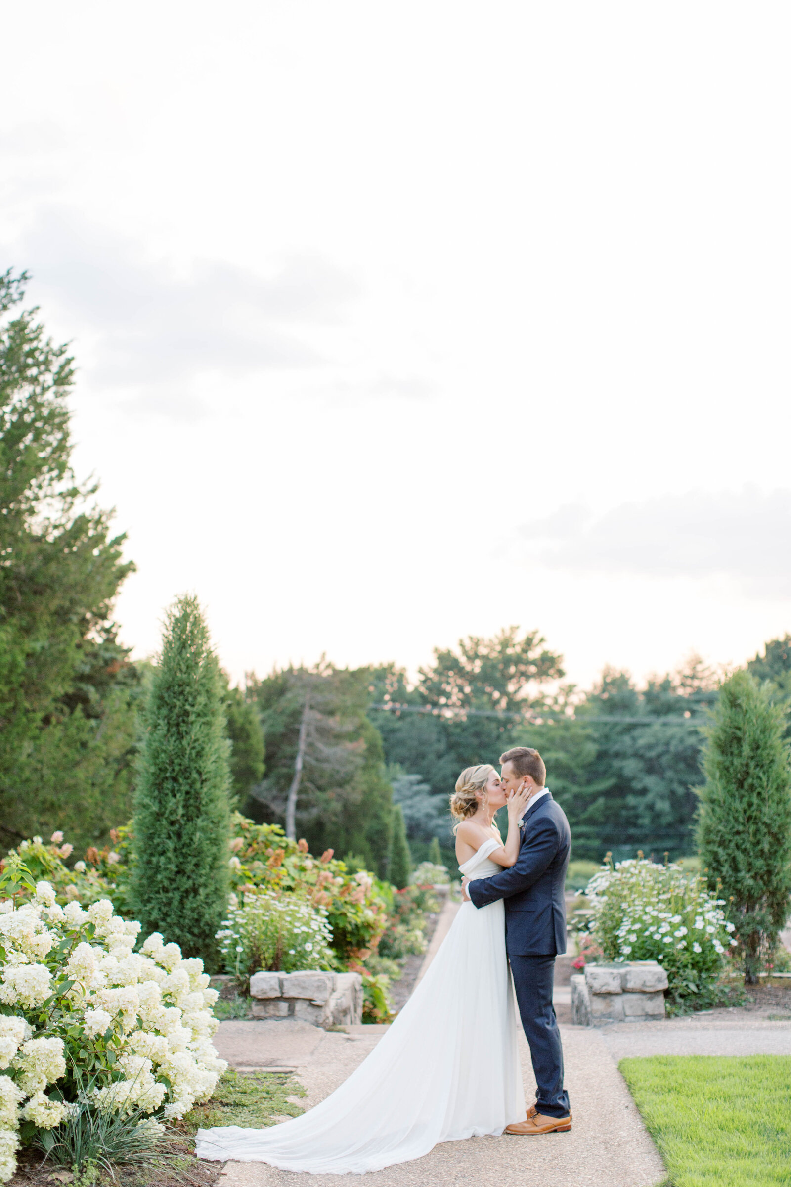 A bride caresses her groom's cheek while kissing him in a garden full of blooms