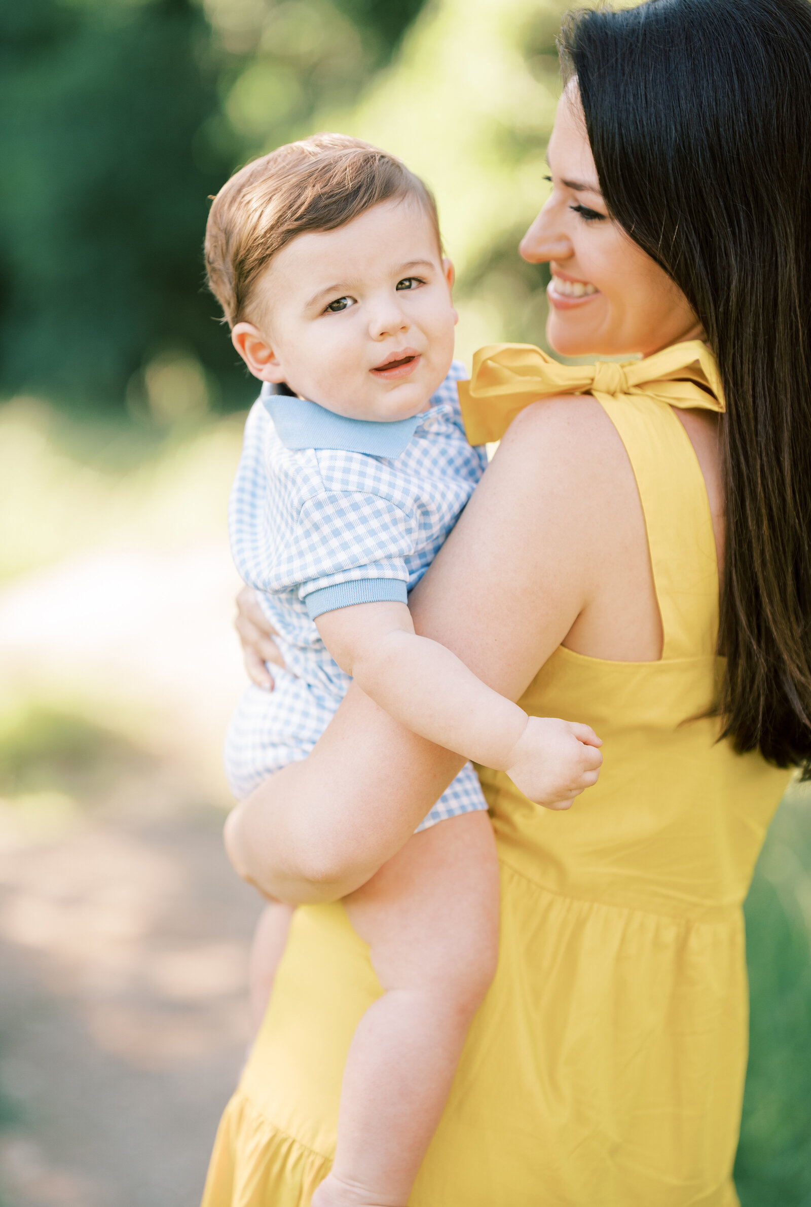 Portrait of a woman in a yellow dress smiling and holding a baby boy on her hip in the outdoors