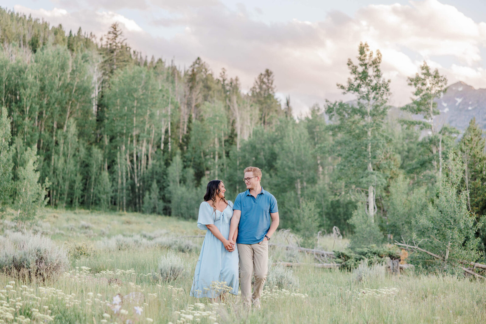 A couple is holding hands and smiling at each other in an aspen filled valley with the mountain peaks behind them