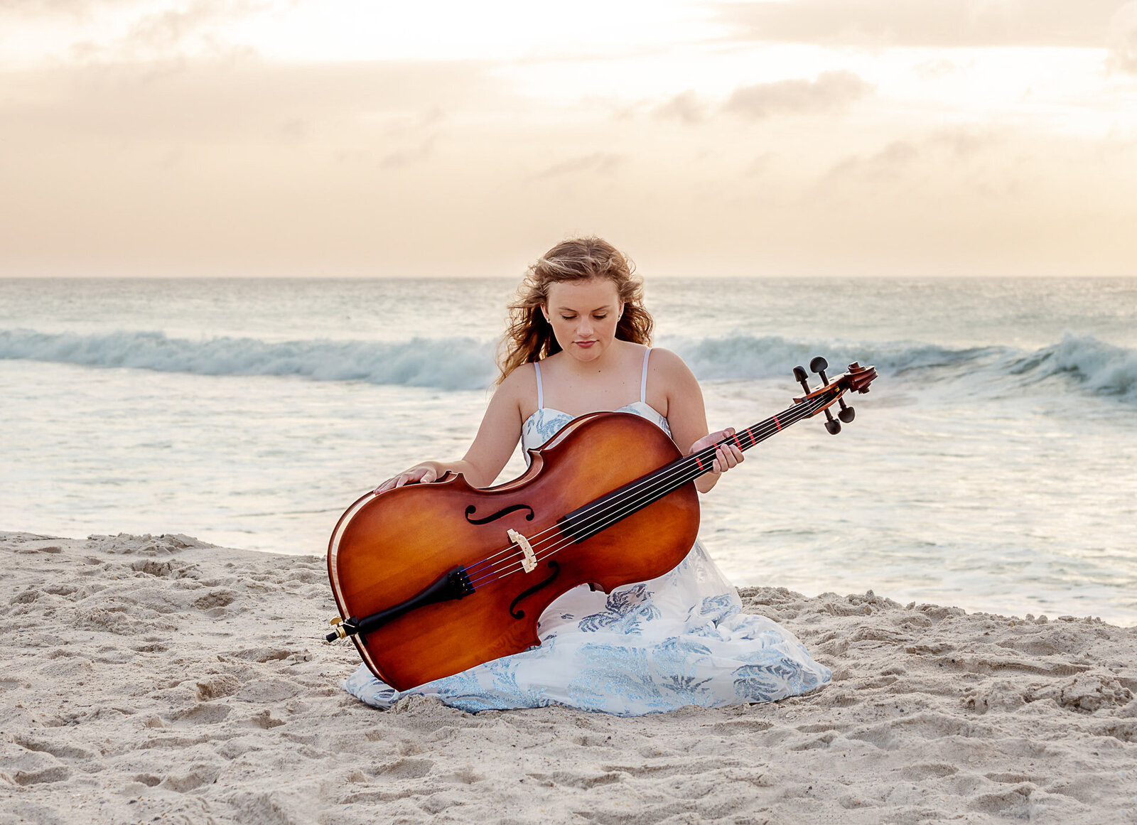 A high school senior in a white dress sitting on the beach with a cello, ocean waves in the background in North Carolina. This artistic photo captures the harmony of music and nature, ideal for those seeking unique senior photography in Wrightsville Beach, NC.