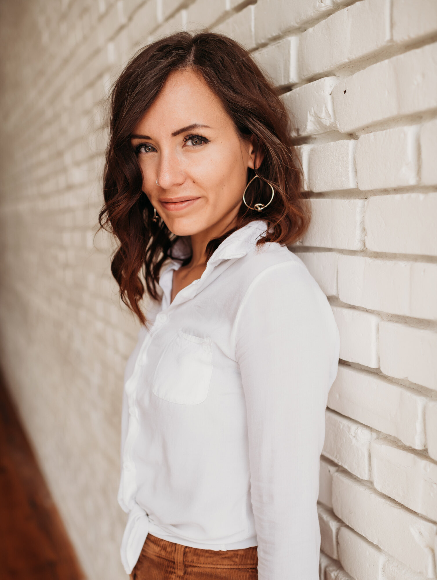 Branding Photographer,  a woman dressed professionally wears a blouse and leans against a white brick wall indoors