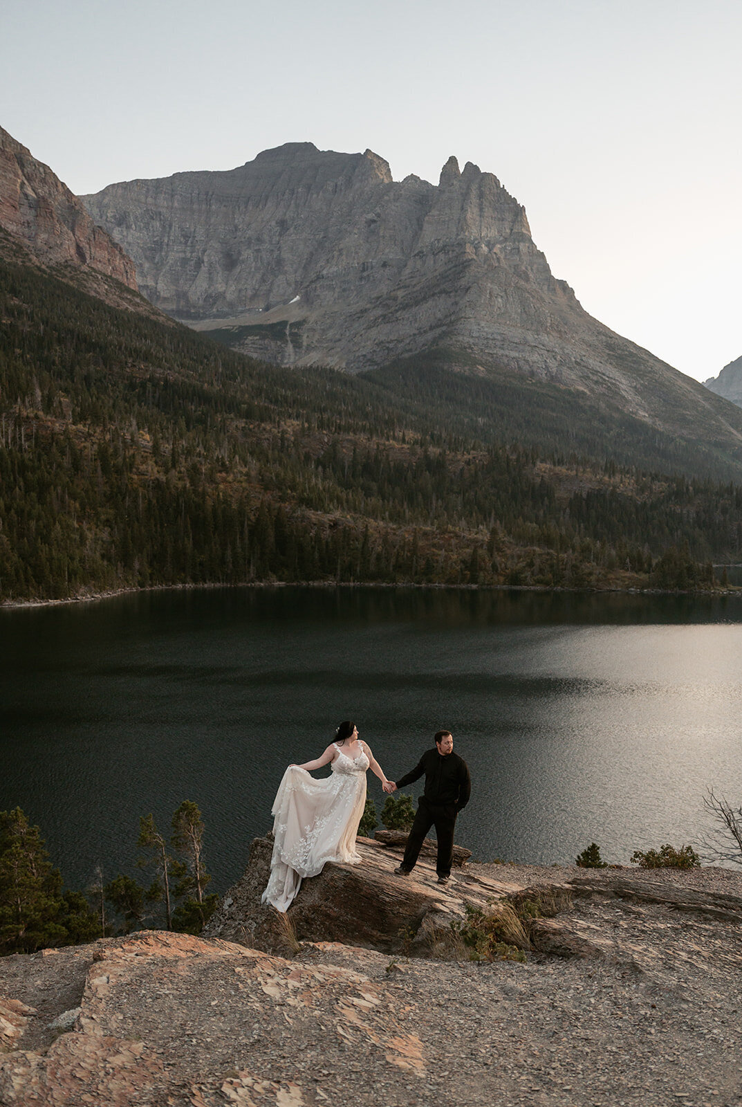 A bride and groom stand on a rocky overlook above a mountain lake surrounded by pine forests and dramatic cliffs during their Glacier National Park elopement, captured by Sydney Breann Photography.