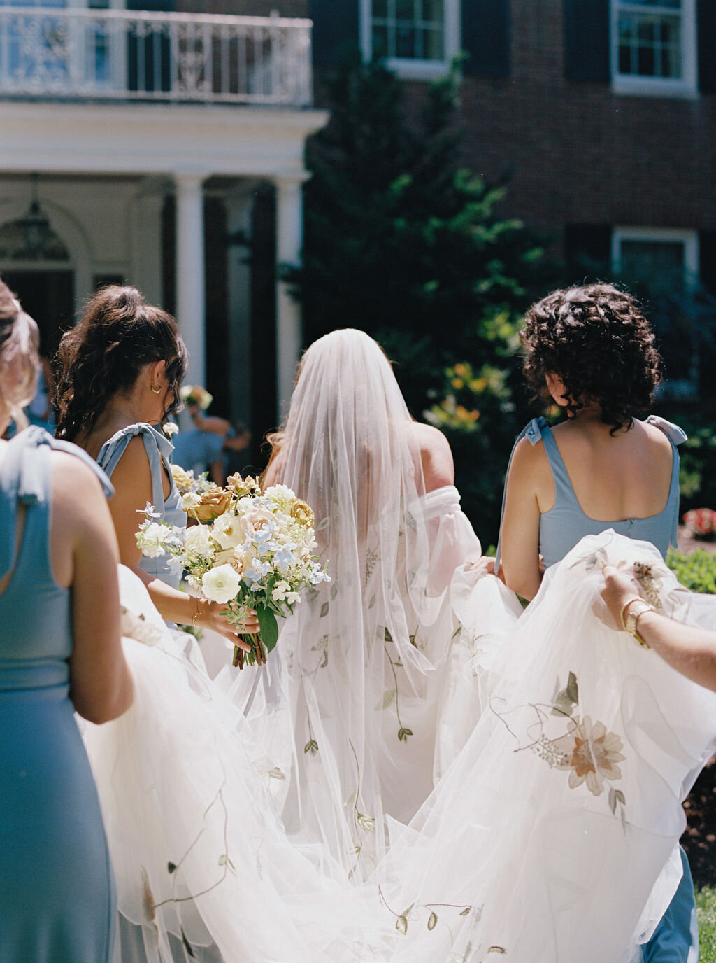 Bridesmaids in blue dresses carrying bride’s flower embroidered veil, train and bridal bouquet.