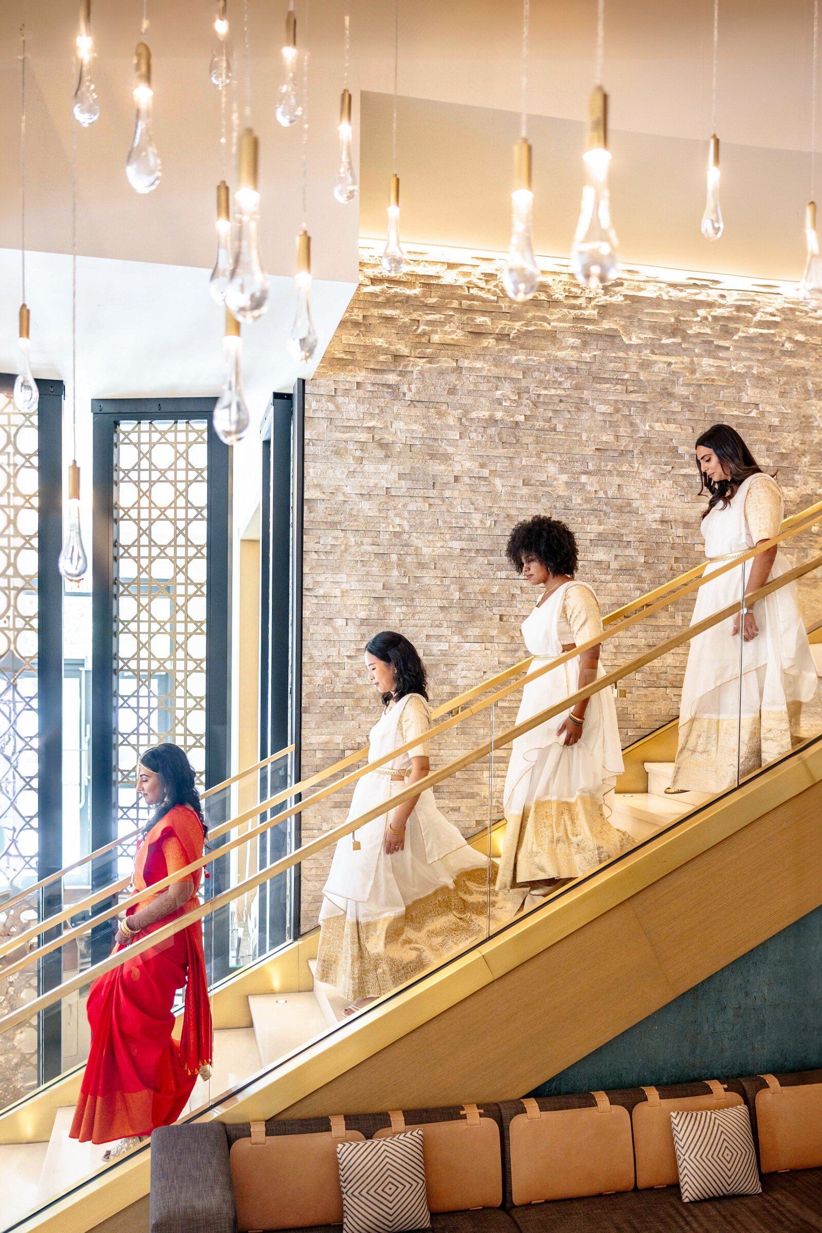 An Indian bride in a red sari leads three women in white dresses down a staircase. The setting features modern decor, with hanging pendant lights and a textured brick wall.