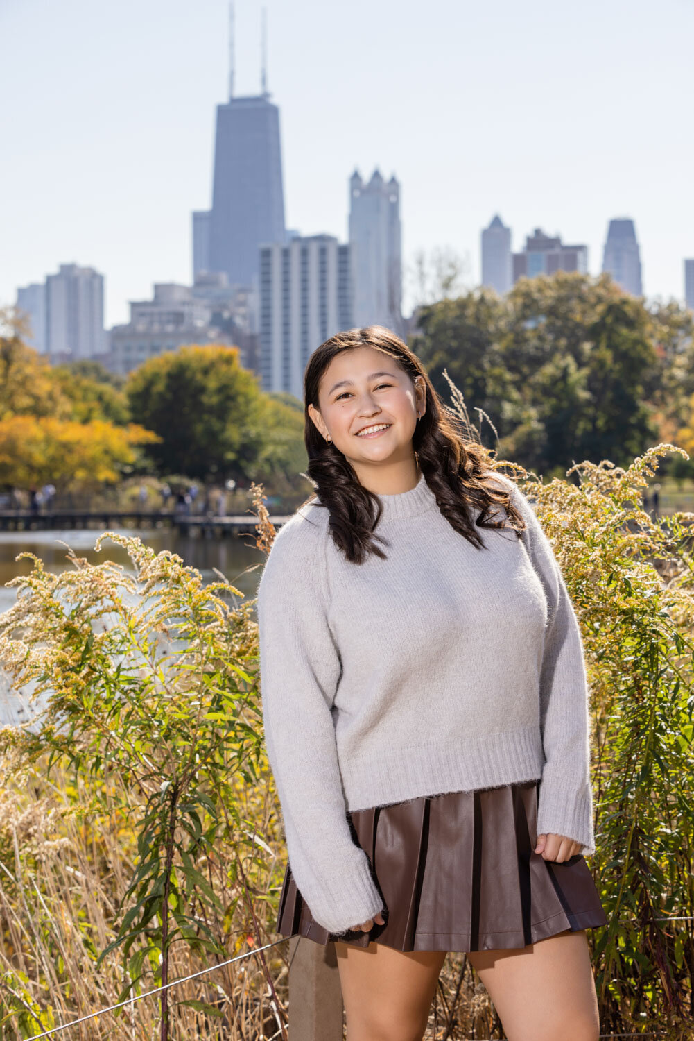 A woman in a grey sweater and brown skirt is standing in front of a city skyline.